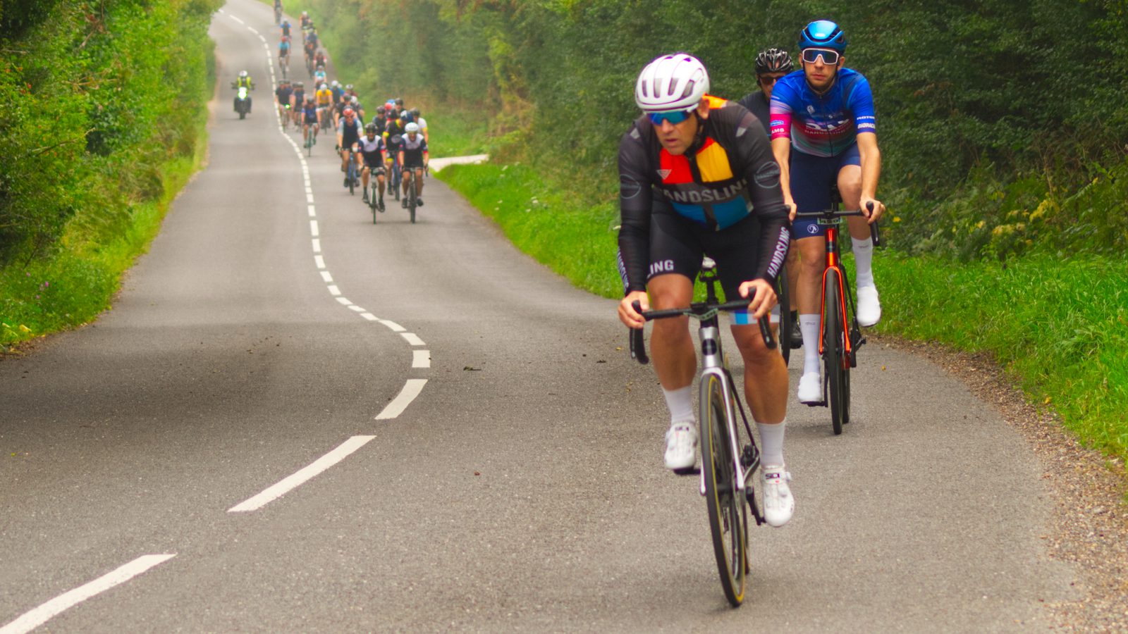 A group of cyclists on a winding country road. The focus is on two cyclists in the forefront, one wearing a white helmet and dark cycling kit, and the other in a blue kit with a blue helmet. Trees and greenery line the road, and others ride behind them.