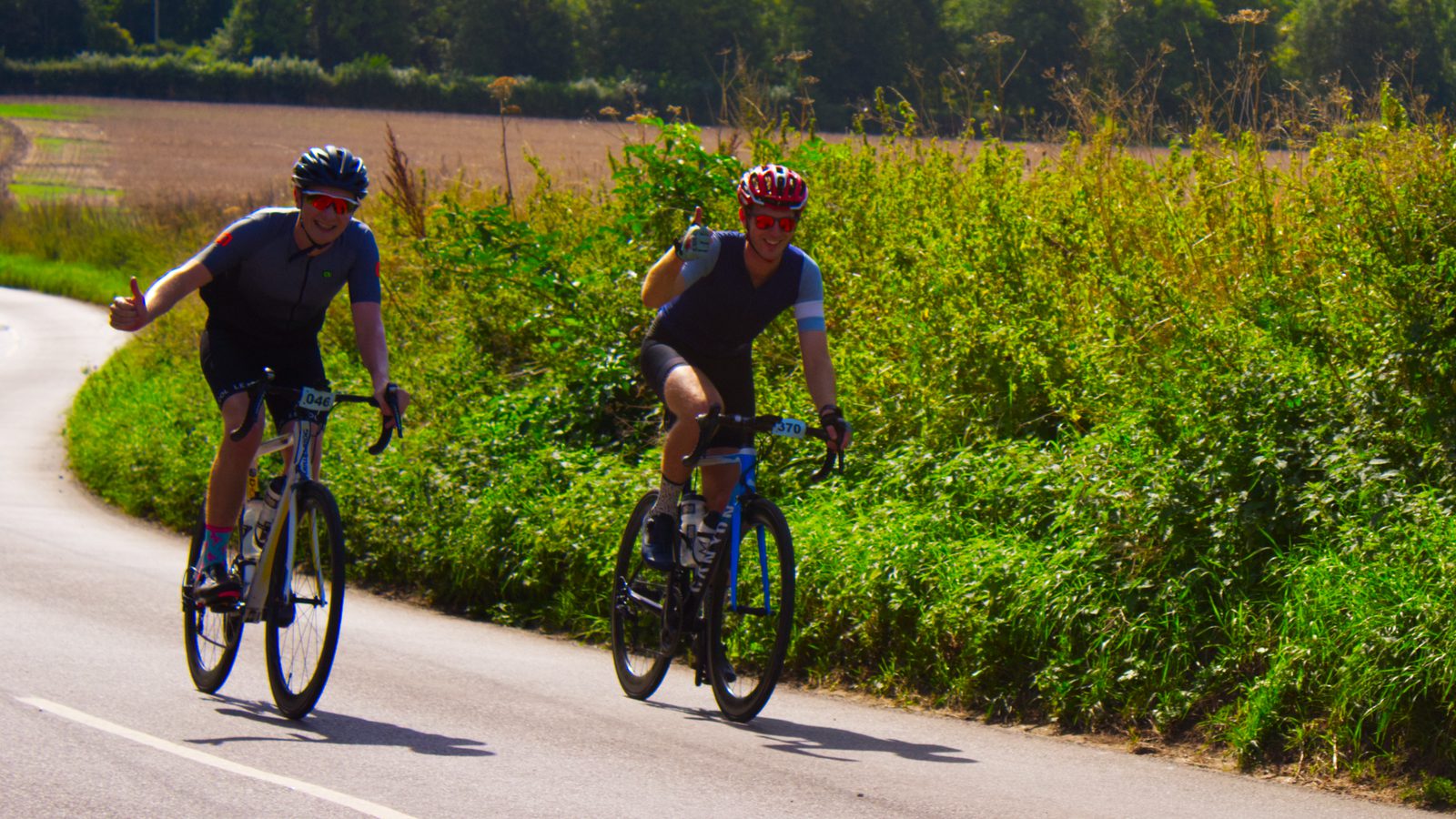 Two bicyclists, wearing helmets and cycling gear, ride along a rural road bordered by lush greenery and fields under a clear sky. One cyclist is waving and smiling, while the other rides close behind, both appearing to enjoy the outdoor activity.