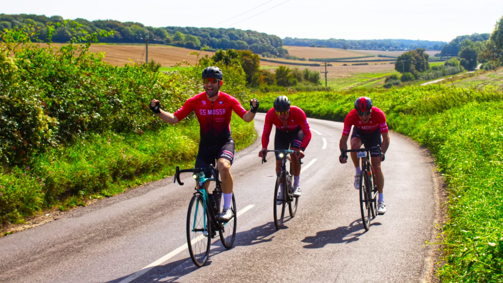 Three cyclists wearing red jerseys ride on a rural road surrounded by lush greenery. The front cyclist raises both arms in celebration, while the other two follow closely behind. The landscape features rolling hills and farmland under a clear, blue sky.
