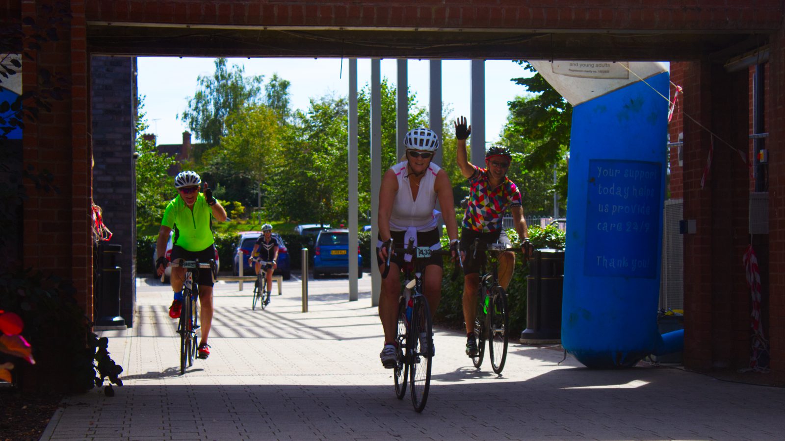 Four cyclists are riding under a brick archway in what appears to be an organized event. The forefront cyclist in white is leading, while a cyclist wearing a colorful shirt waves. The background shows greenery and additional buildings under a bright sky.