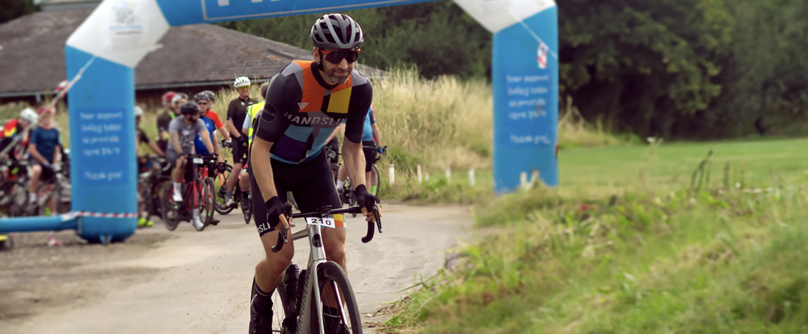 A cyclist in a colorful jersey and black shorts rides along a path, smiling, with a race number "210" on his bike. Behind him, a group of cyclists gather at the starting point under an inflatable arch. The background includes trees and grass.