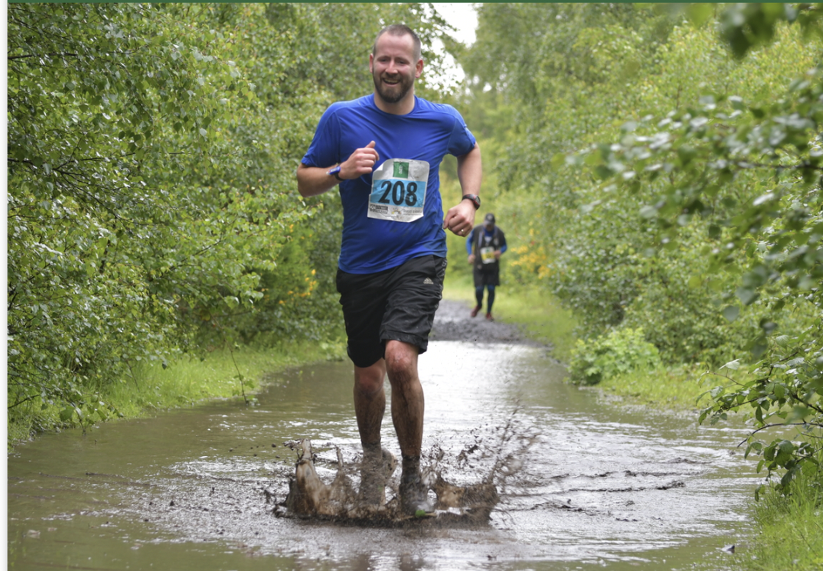 A man in a blue shirt and black shorts, with a race bib numbered 208, is running through a muddy puddle on a forest trail. He is smiling and splashing water. Another runner can be seen in the distance. The trail is surrounded by lush green trees and vegetation.