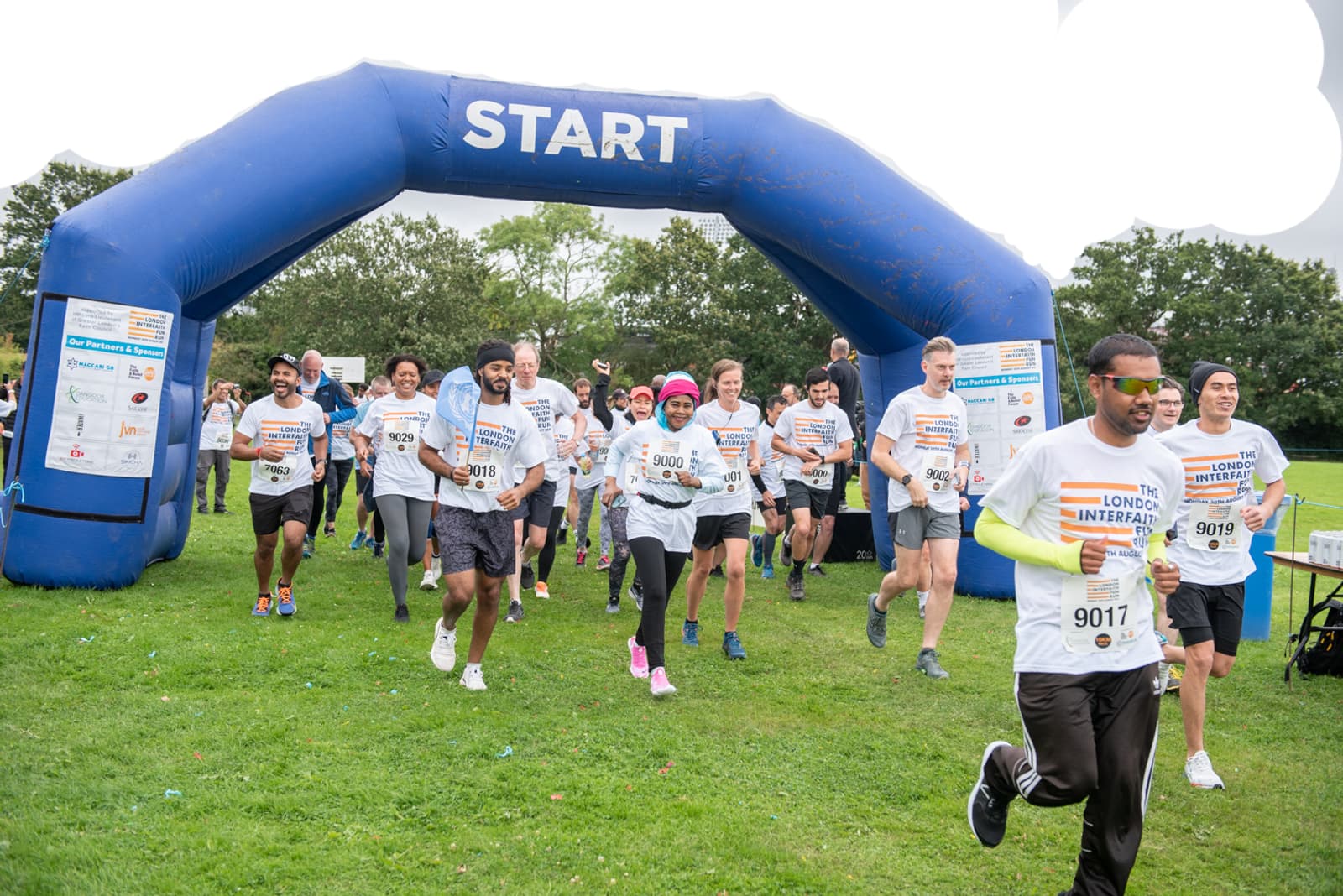 A diverse group of runners, wearing numbered bibs, start a race by passing under a blue inflatable arch with "START" written on it. The participants are smiling and dressed in various athletic attire. The event is taking place on a grassy area with trees in the background.