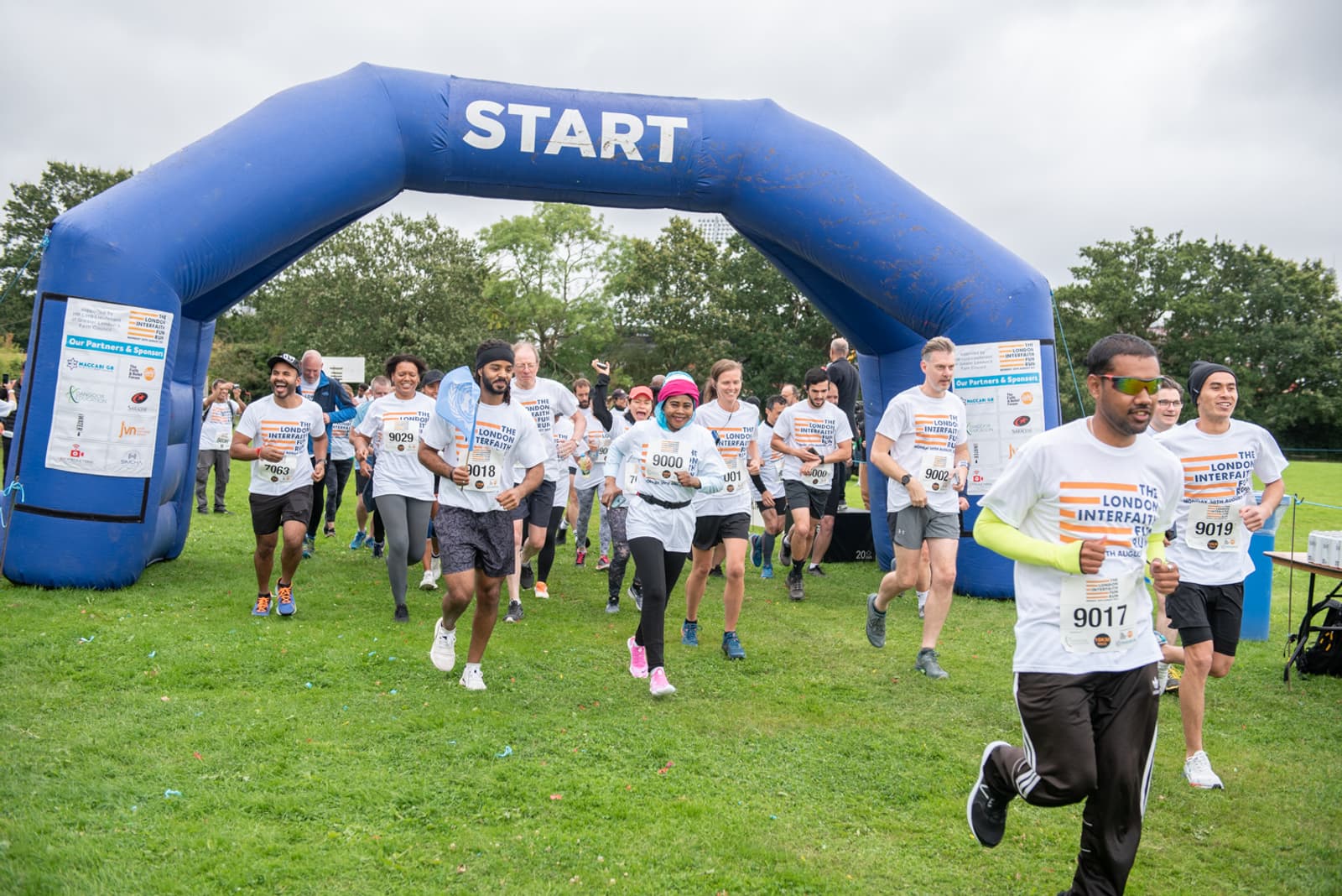 Participants in a community charity run are starting the race, passing under a large blue "START" arch. The runners, wearing numbered bibs and event T-shirts, are on a grassy field, smiling and enthusiastic. The sky is overcast, and trees are in the background.