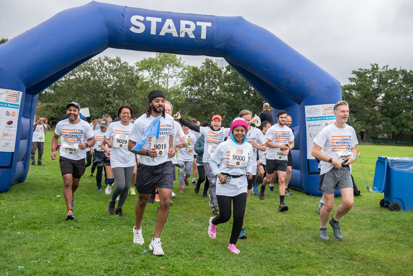 A group of runners in athletic attire participates in an outdoor charity race, smiling as they pass under a blue inflatable start arch. The participants wear event bibs, and the sky is overcast with trees and grassy surroundings in the background.