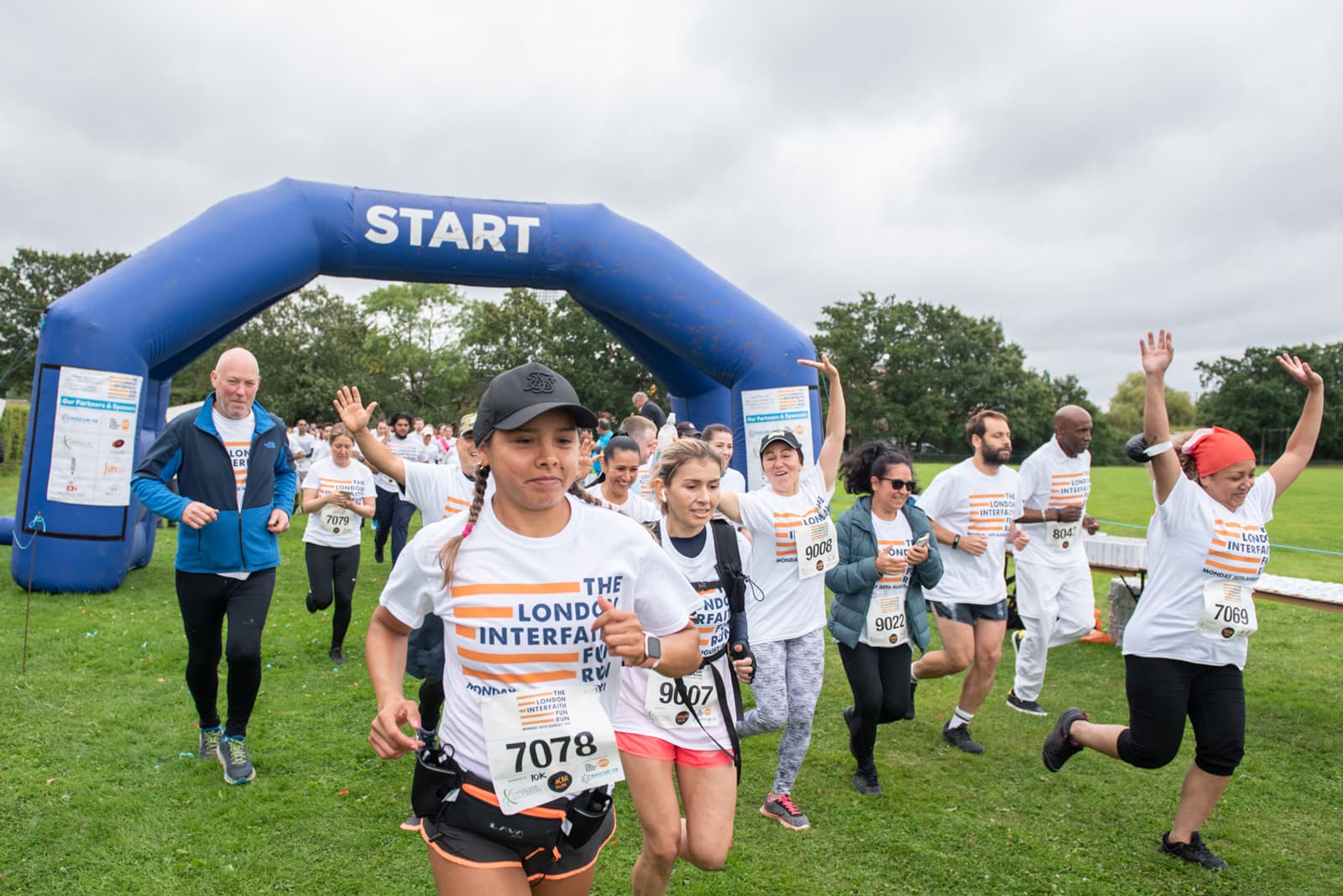 A diverse group of runners starting a race under a blue inflatable arch marked "START." They are wearing white T-shirts with "The London Interfaith Fun Run" printed on them. The participants display enthusiasm, with some raising their arms and smiling.