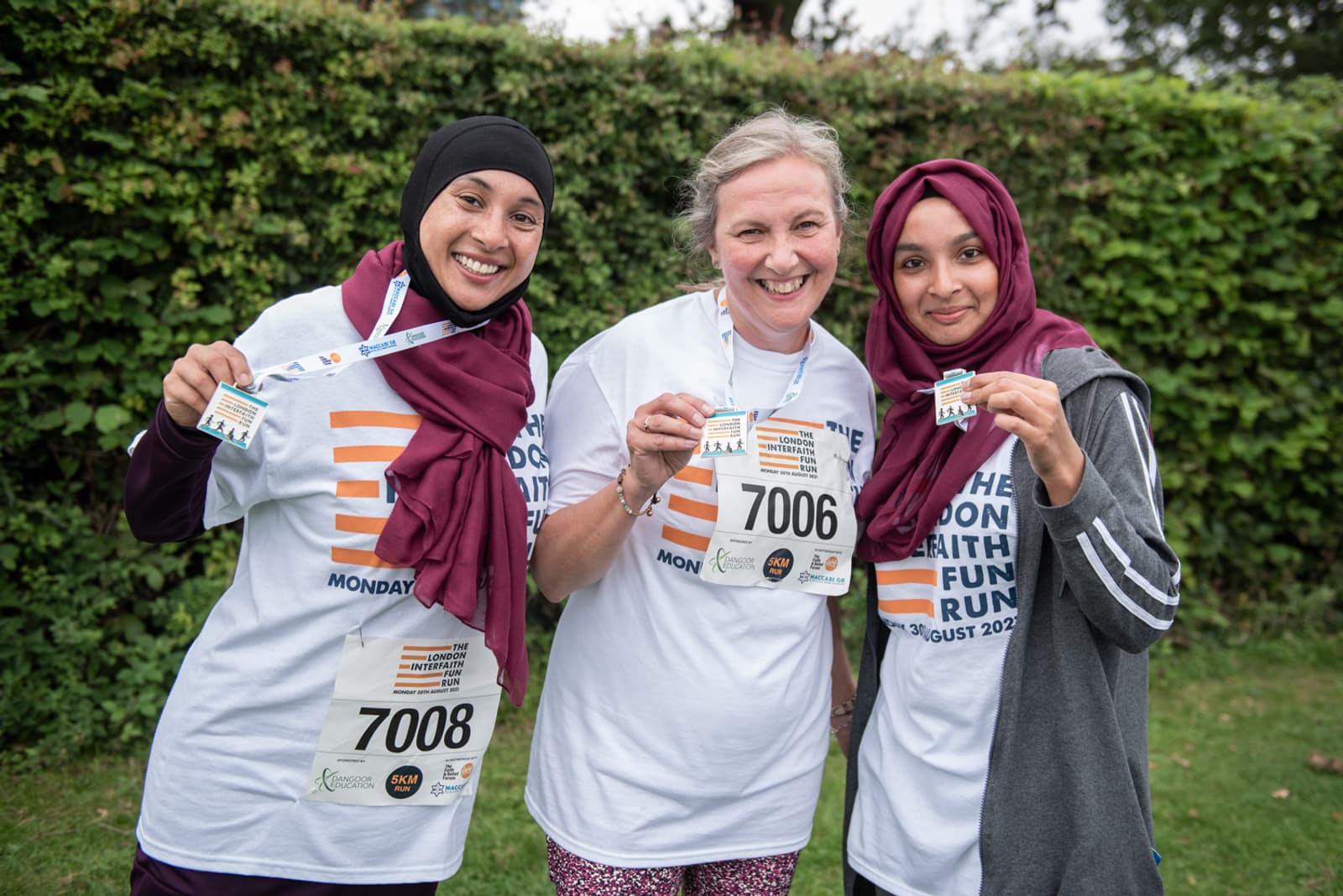 Three smiling women dressed in matching white t-shirts and holding medals pose for a photo at a fun run event. They are outdoors with green foliage in the background. Two are wearing headscarves, and all of their race bibs are visible.