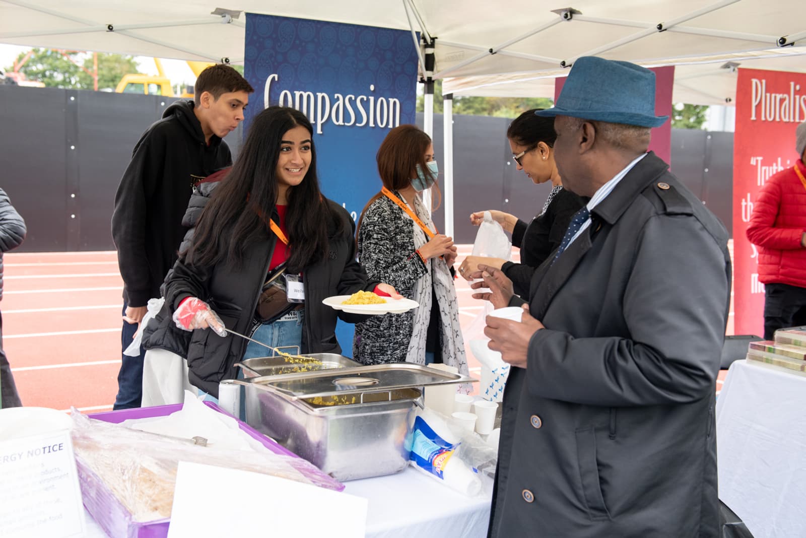 People are gathered in a tent at a community event. One person is serving food to another, and there are banners with words like "Compassion" and "Pluralism" in the background. Food trays and supplies are visible on the table.