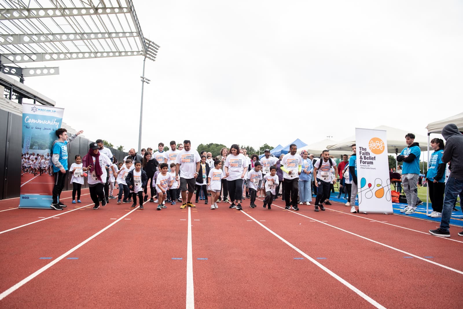 A diverse group of people, including children, line up at the starting line of a race on a red track. They are about to start running. Volunteers and spectators stand nearby, with banners and tents visible in the background on a cloudy day.