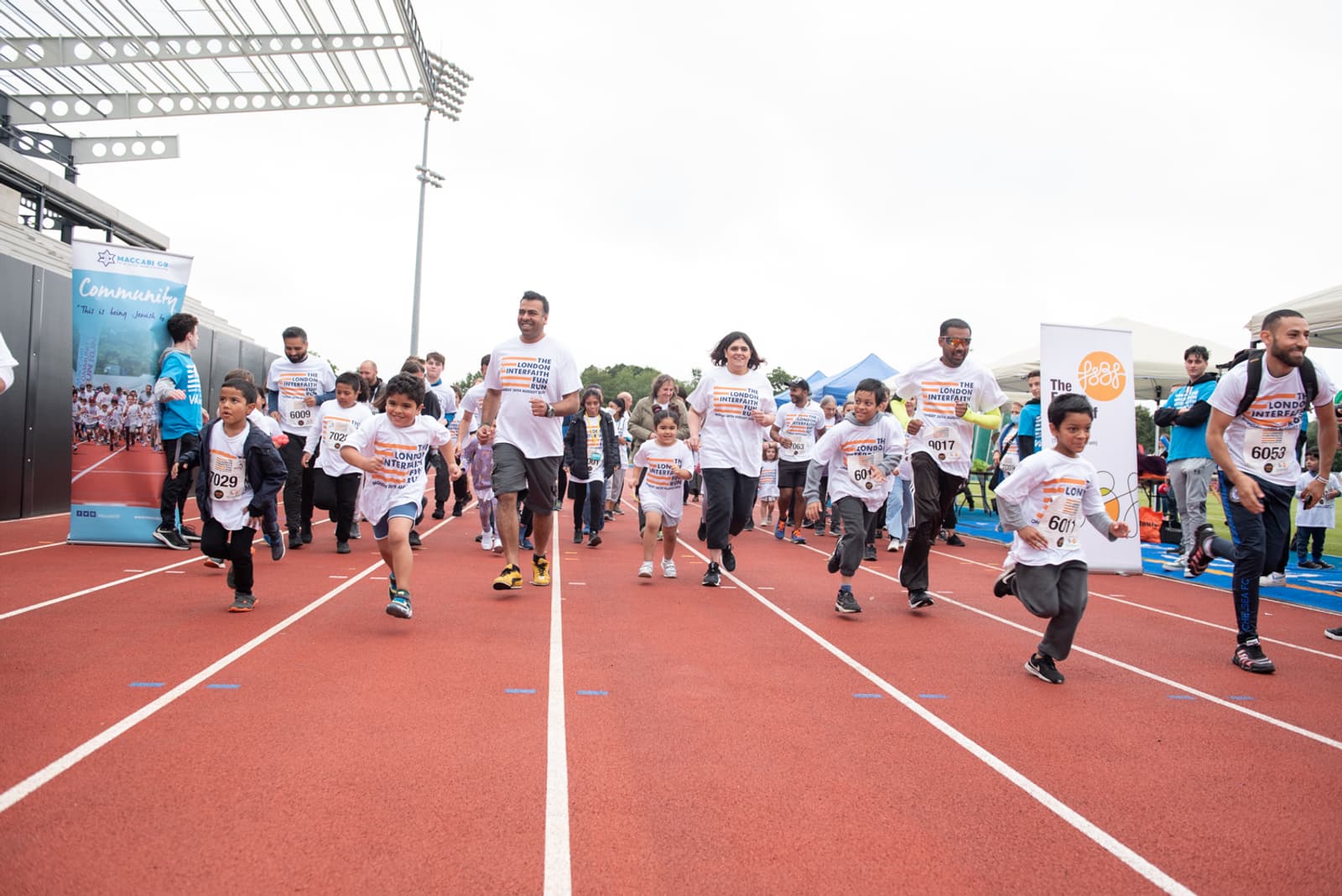 A group of people of various ages, including children and adults, are running on a track during an organized event. They are wearing matching white T-shirts with numbers pinned on them. The background includes a banner and a few tents.