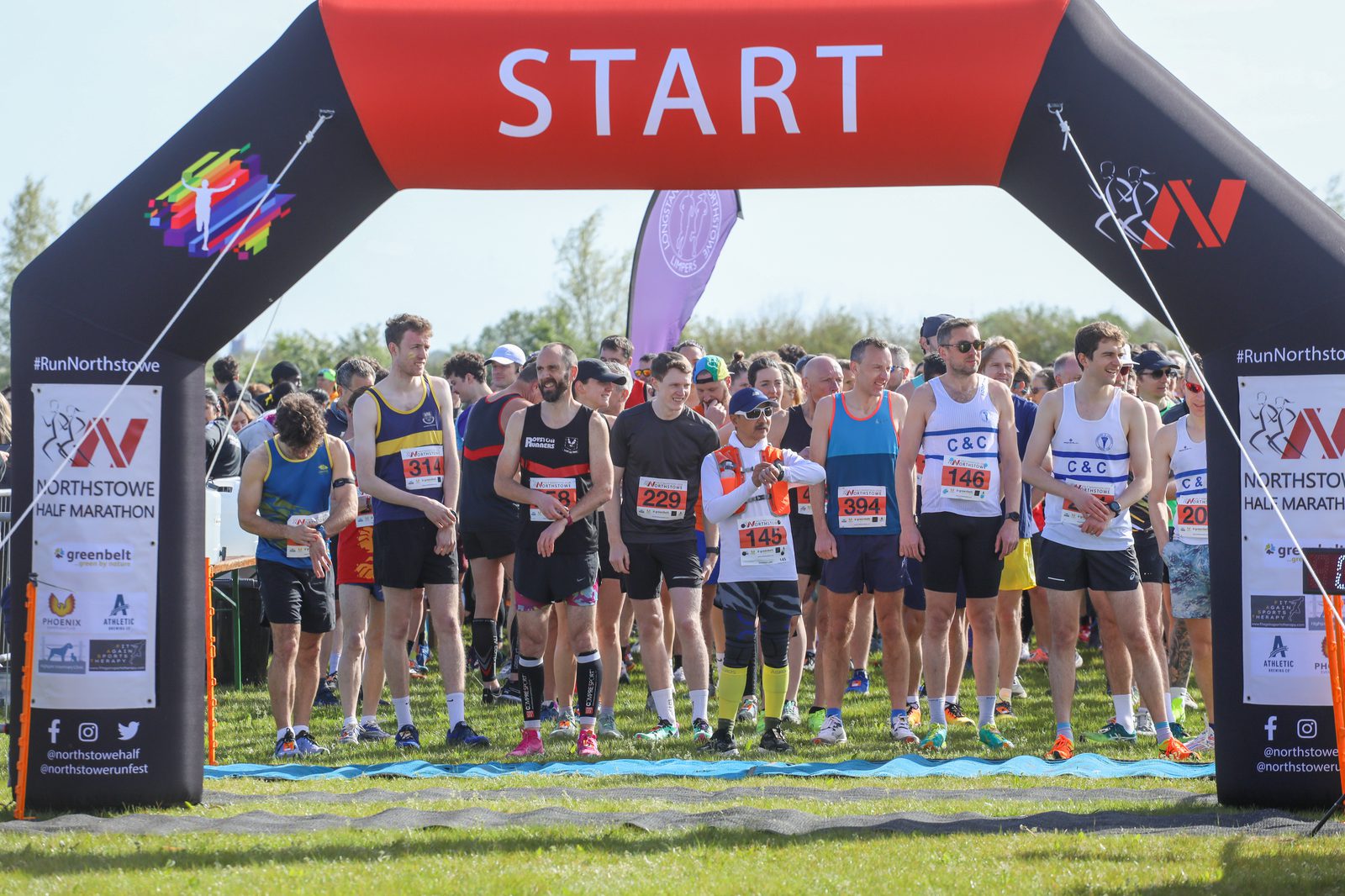 A group of runners is lined up at the start line of a race under a large black and red inflatable archway with the word "START" printed on it. The runners are wearing various running gear and numbered bibs, preparing to begin the race on a clear, sunny day.