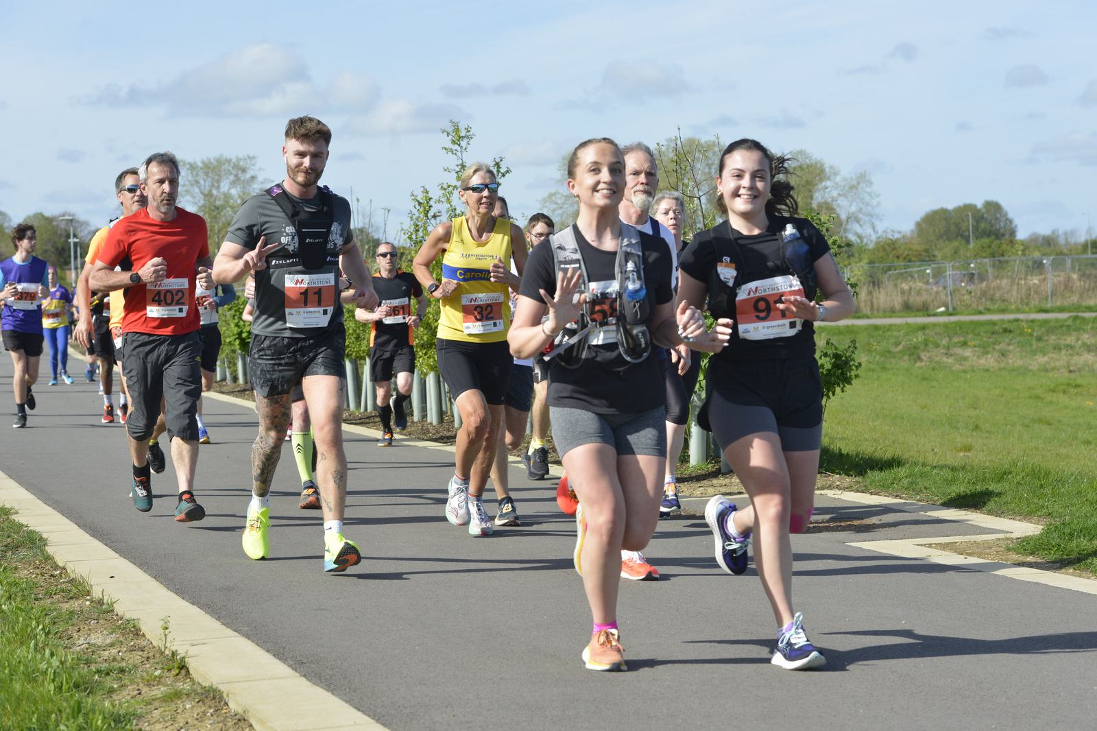 A group of marathon runners on a sunny day, smiling and jogging on a paved path surrounded by greenery. The participants have race numbers pinned to their shirts. The atmosphere is energetic and cheerful, with diverse individuals running together.