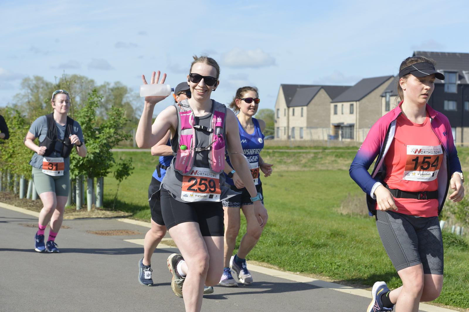 Runners participate in a race on a sunny day. One woman waves with a water bottle in hand, wearing a pink hydration vest and sunglasses. Others run alongside her, all wearing race bibs. Houses and trees line the background.
