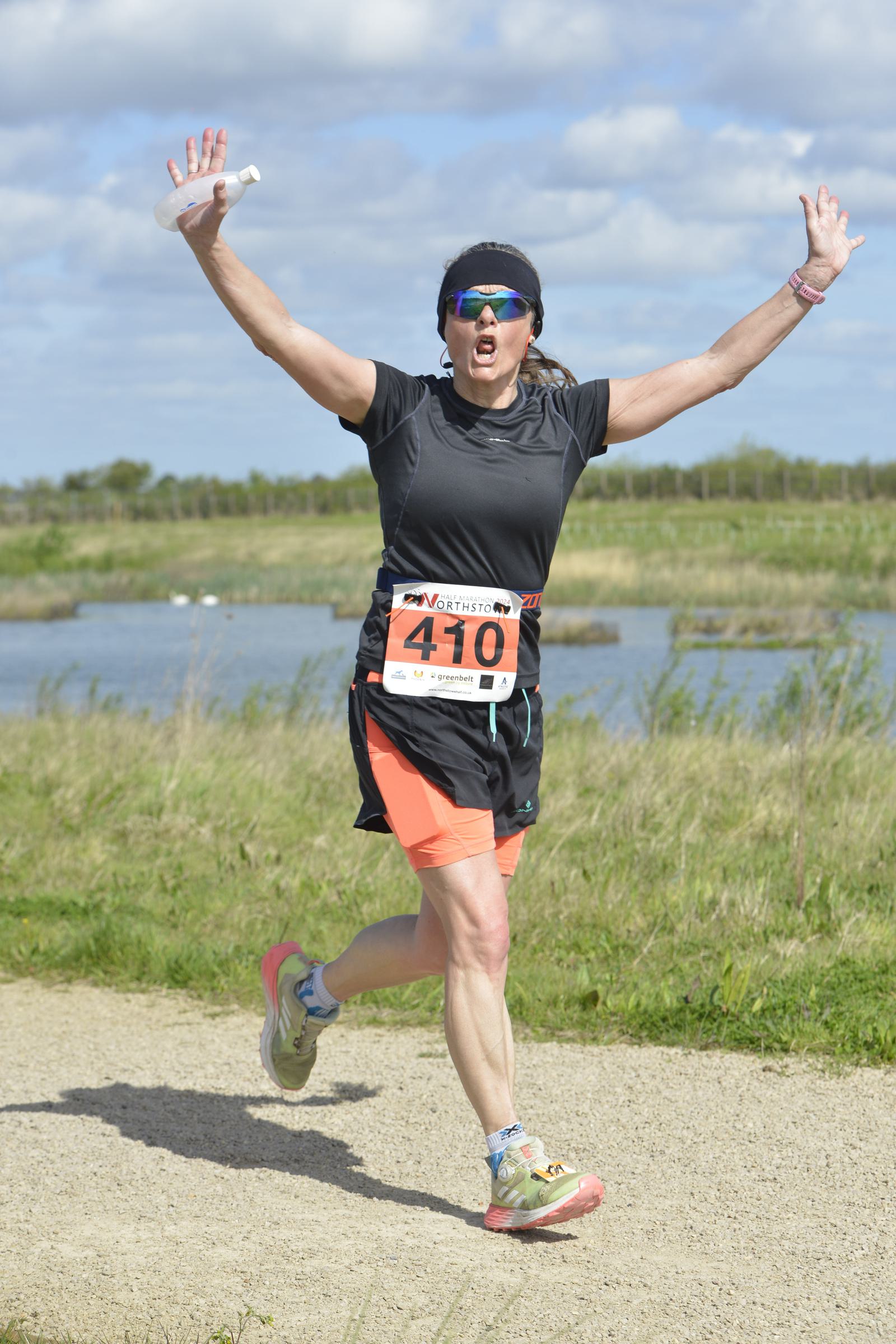 A person wearing athletic gear and sunglasses is joyfully running on a gravel path, with arms raised in triumph. They wear a race bib with the number 410. The background features grass, water, and a partly cloudy sky.