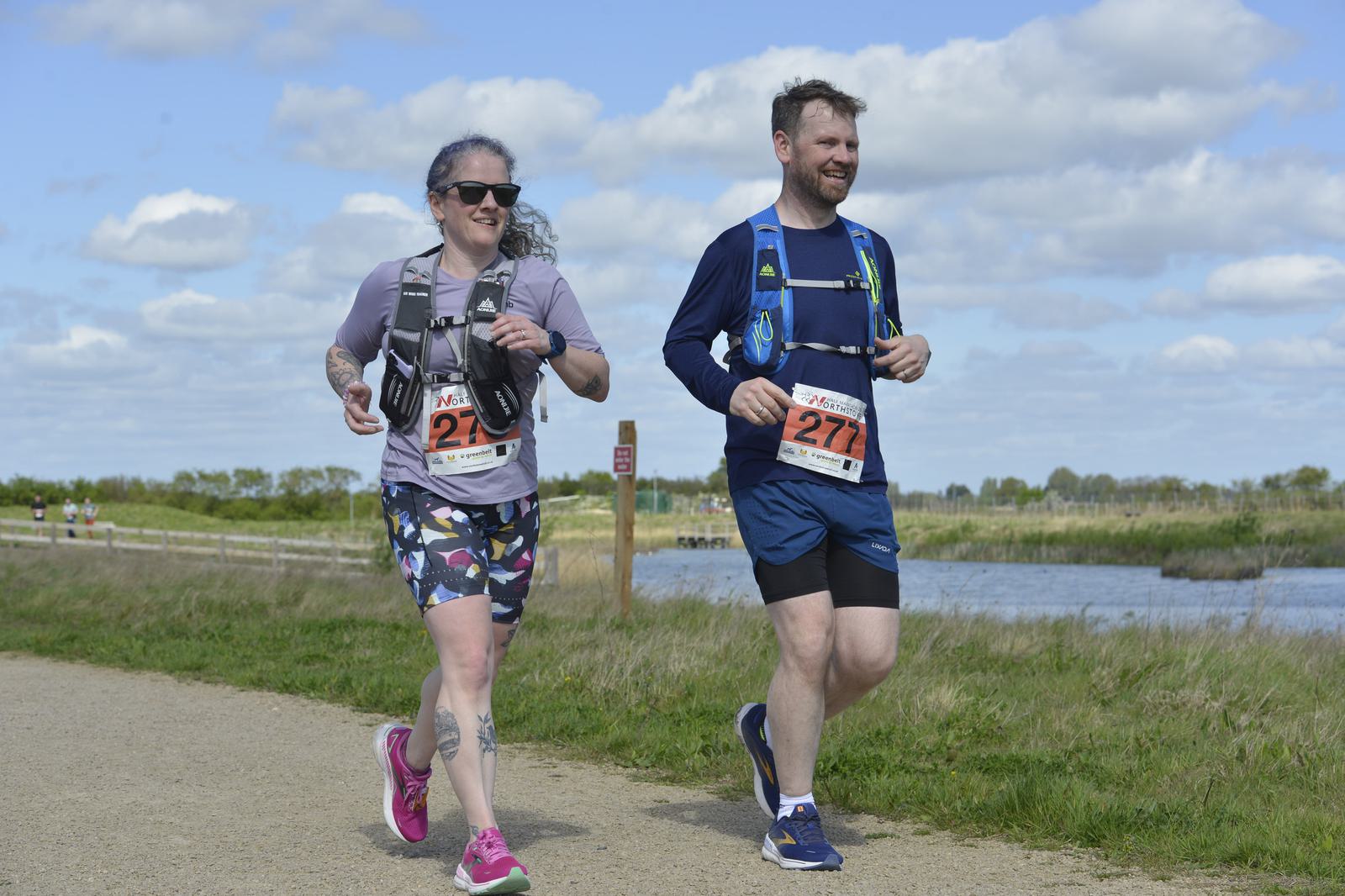 Two people are jogging on a pathway near a river. The woman on the left wears a gray shirt and colorful shorts, while the man on the right wears a blue shirt and black shorts. They both have race numbers and hydration packs. It's a sunny day with clouds.