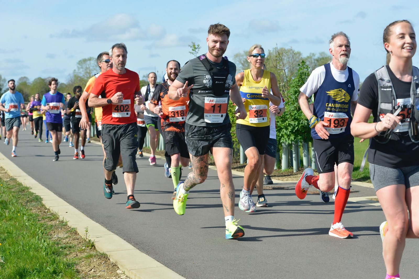 A group of runners participates in a race on a sunny day. The diverse group includes men and women of various ages, dressed in athletic gear and numbered bibs, running along a paved path surrounded by greenery. Some runners smile and wave at the camera.