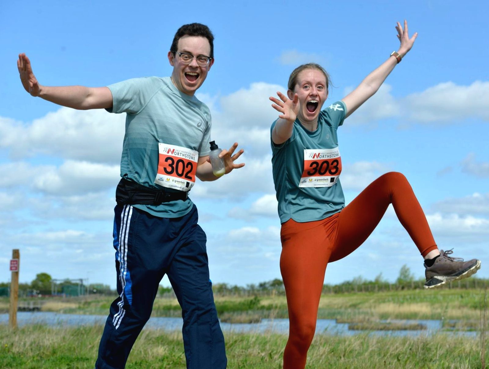 Two people in athletic clothing and race bibs (302 and 303) enthusiastically pose for the camera with big smiles and expressive gestures. They are outdoors on a sunny day with a grassy field, a lake, and a blue sky with scattered clouds in the background.