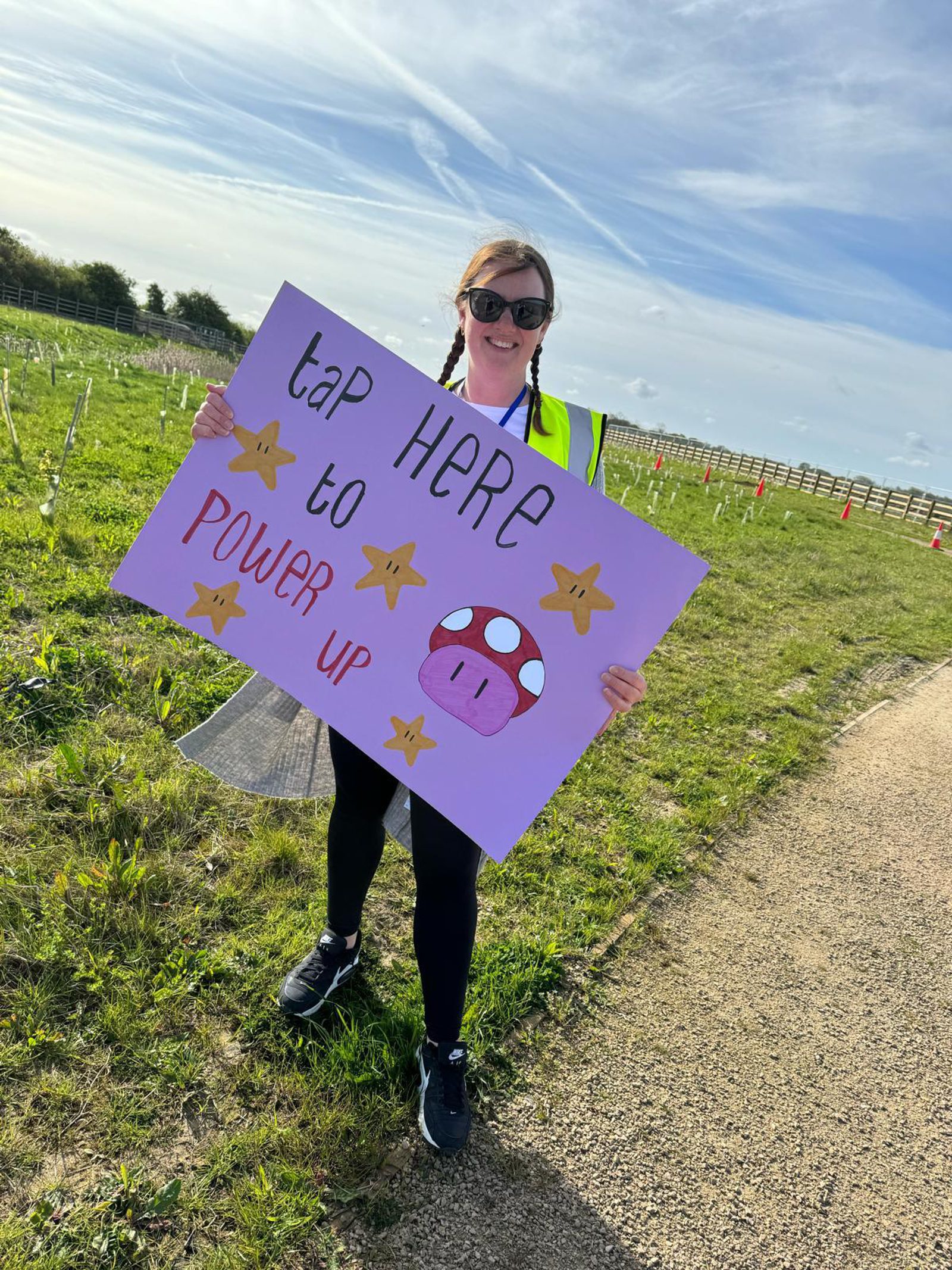 A woman in a high-visibility vest stands on a path in a grassy area under a blue sky with scattered clouds. She holds a pink sign that reads "tap HERE to POWER up" with stars and a red mushroom with white spots from the Mario series as decorations.