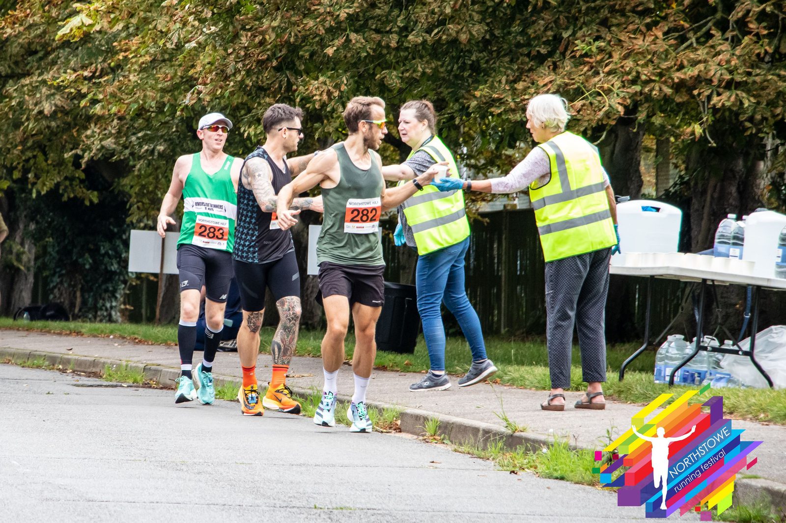 Three runners wearing race bibs numbered 283, 281, and 282, pass by a hydration station during a road race. Two volunteers in yellow vests offer them water. The backdrop includes trees and a table with refreshments. A colorful logo is visible in the bottom right corner.
