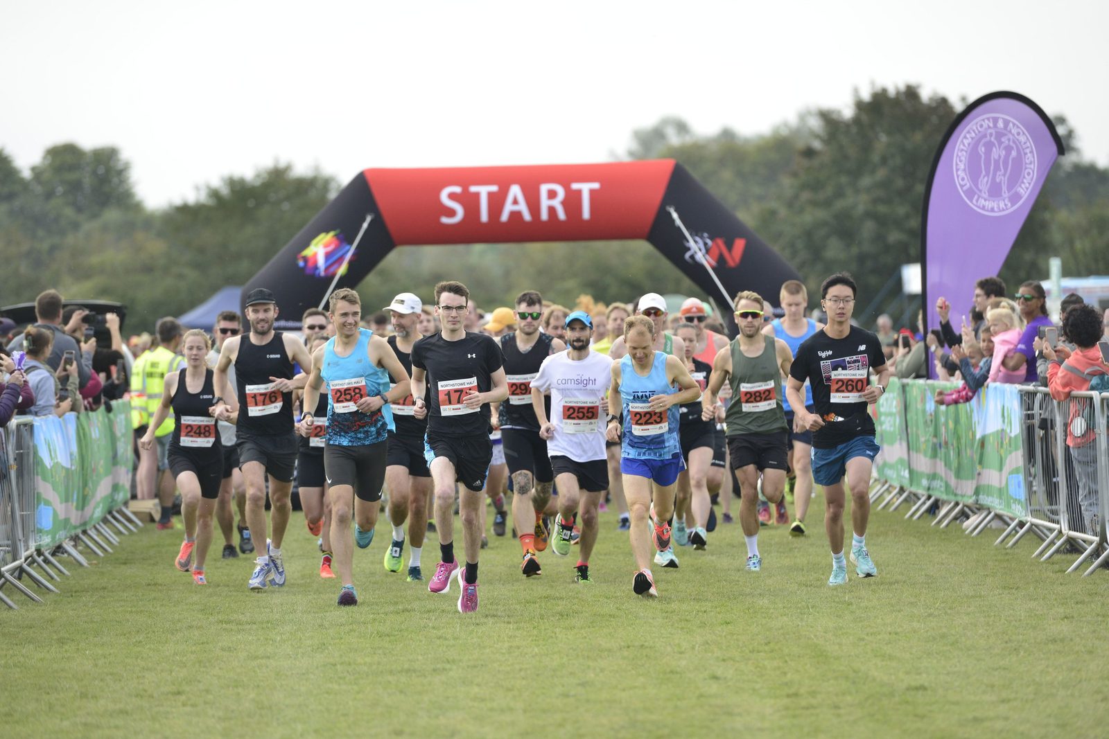 A group of runners, wearing athletic gear and numbered bibs, are starting a race on a grassy field. A large red inflatable arch with the word "START" in white letters is visible in the background. Spectators and banners line the course. Trees are in the distance.