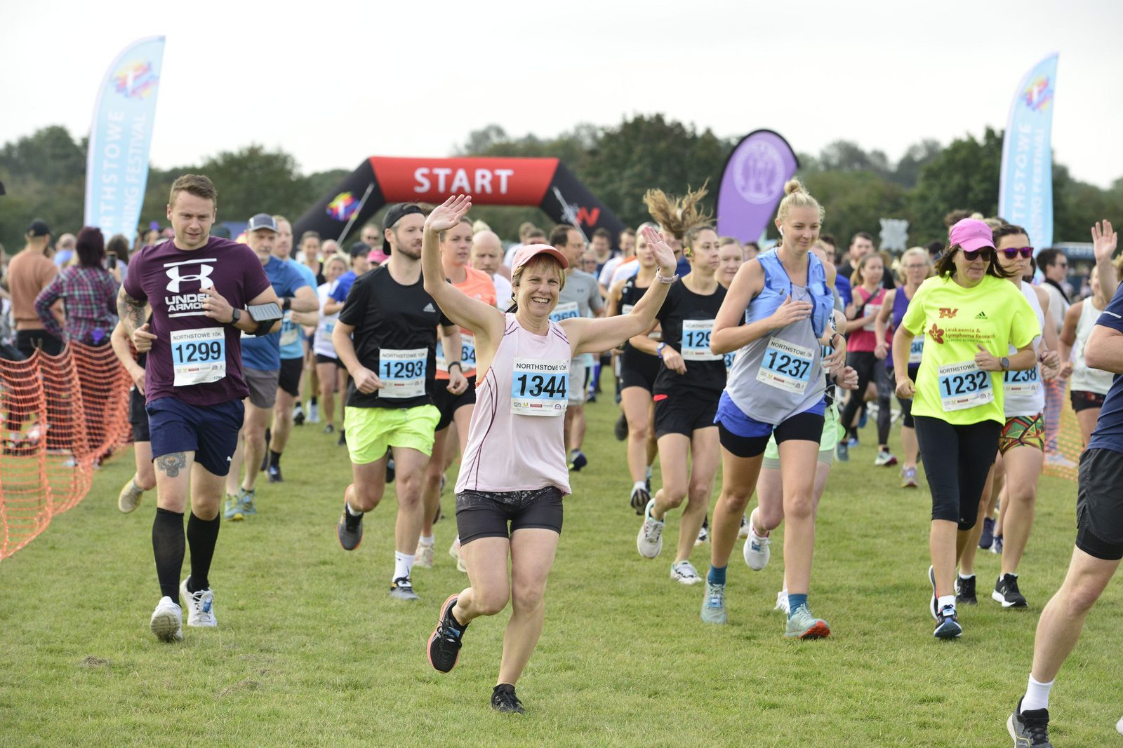 A group of runners, including a woman in a pink cap and bib number 1344 raising her hands, are participating in a race. They are on a grassy field, with a red "START" arch and spectators in the background. Other runners wear various athletic clothing and bib numbers.