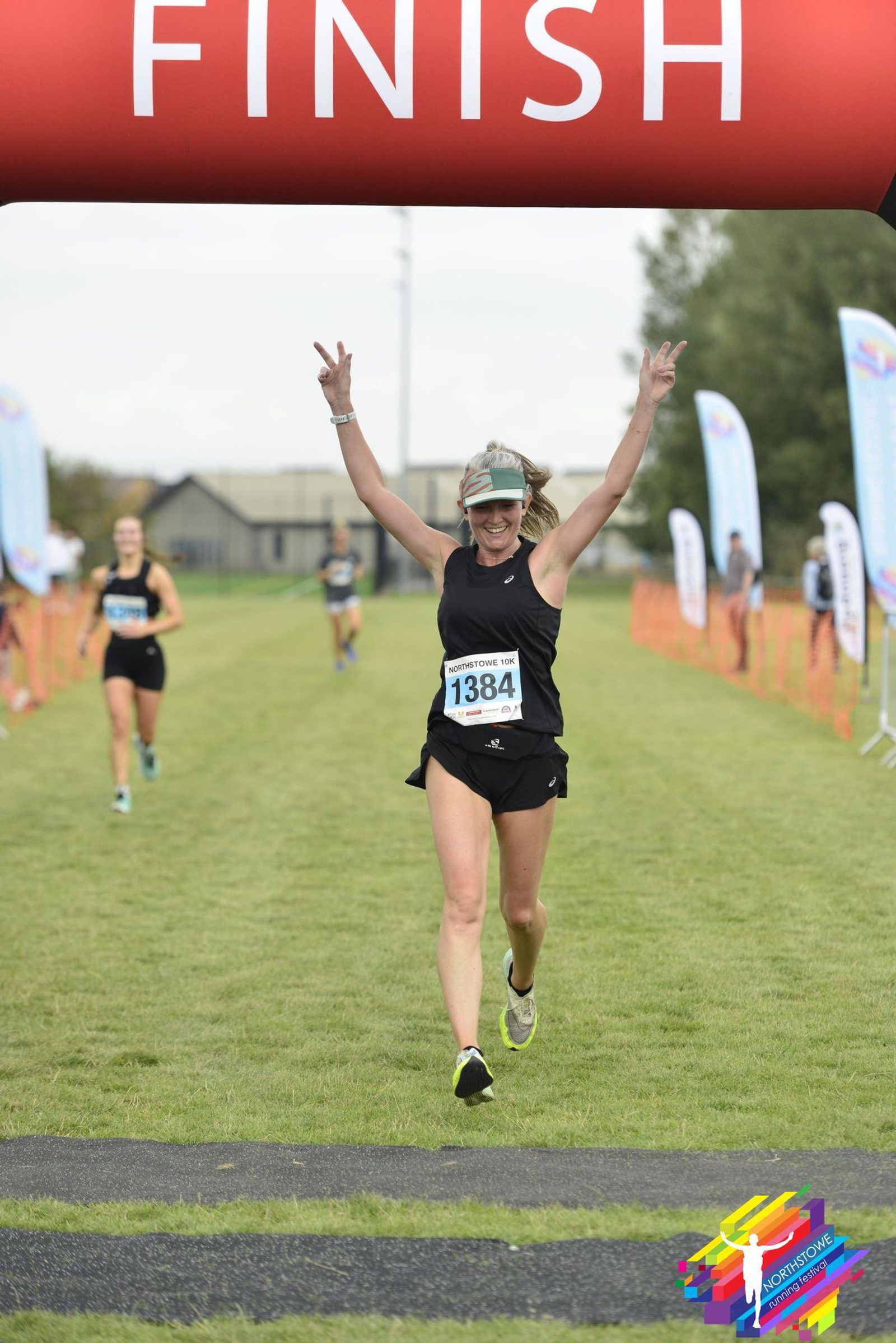 A runner, wearing a black outfit and numbered bib 1384, joyfully crosses the finish line with both arms raised. The finish line banner is red with white letters. Other runners, race flags, and blurred spectators are visible in the background.