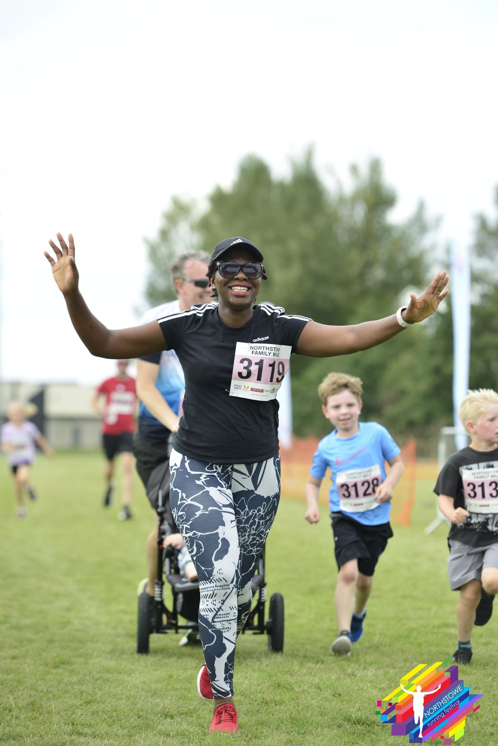 A smiling woman wearing a race bib, sunglasses, and patterned leggings is running with her arms raised. Several children, also with race bibs, are running alongside her. Behind them, a man is pushing a stroller on a grassy field. Trees and a banner are in the background.
