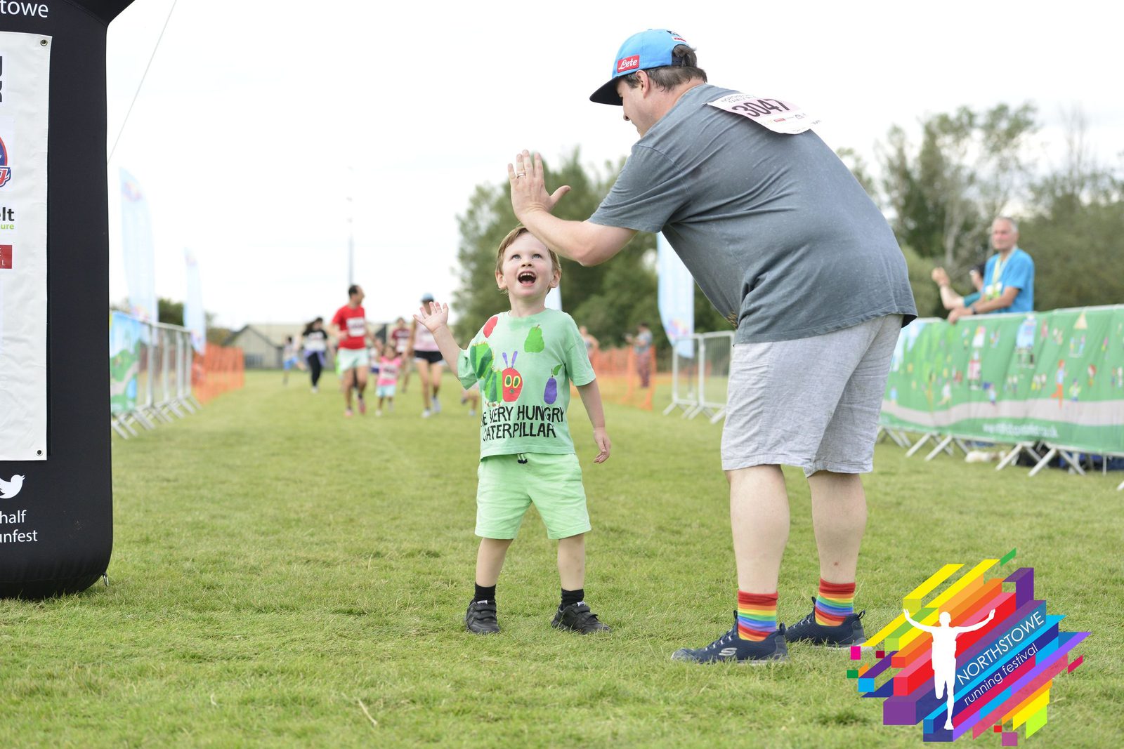 A man and a young boy wearing green, both standing on a grassy field, share a high five at a running event. Other participants and some event banners can be seen in the background, and the boy looks thrilled. The man wears a gray shirt, gray shorts, and rainbow socks.