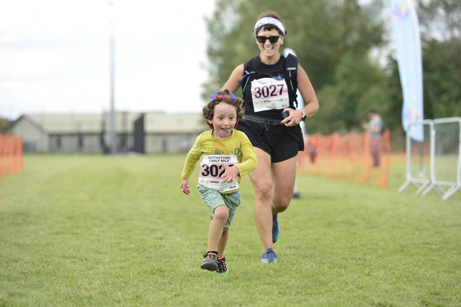 A smiling child wearing a yellow shirt and green shorts runs on grass with a race bib numbered 3027, closely followed by a smiling adult in athletic gear with a race bib numbered 3027. Orange safety netting lines the path, and other racers and onlookers are in the background.