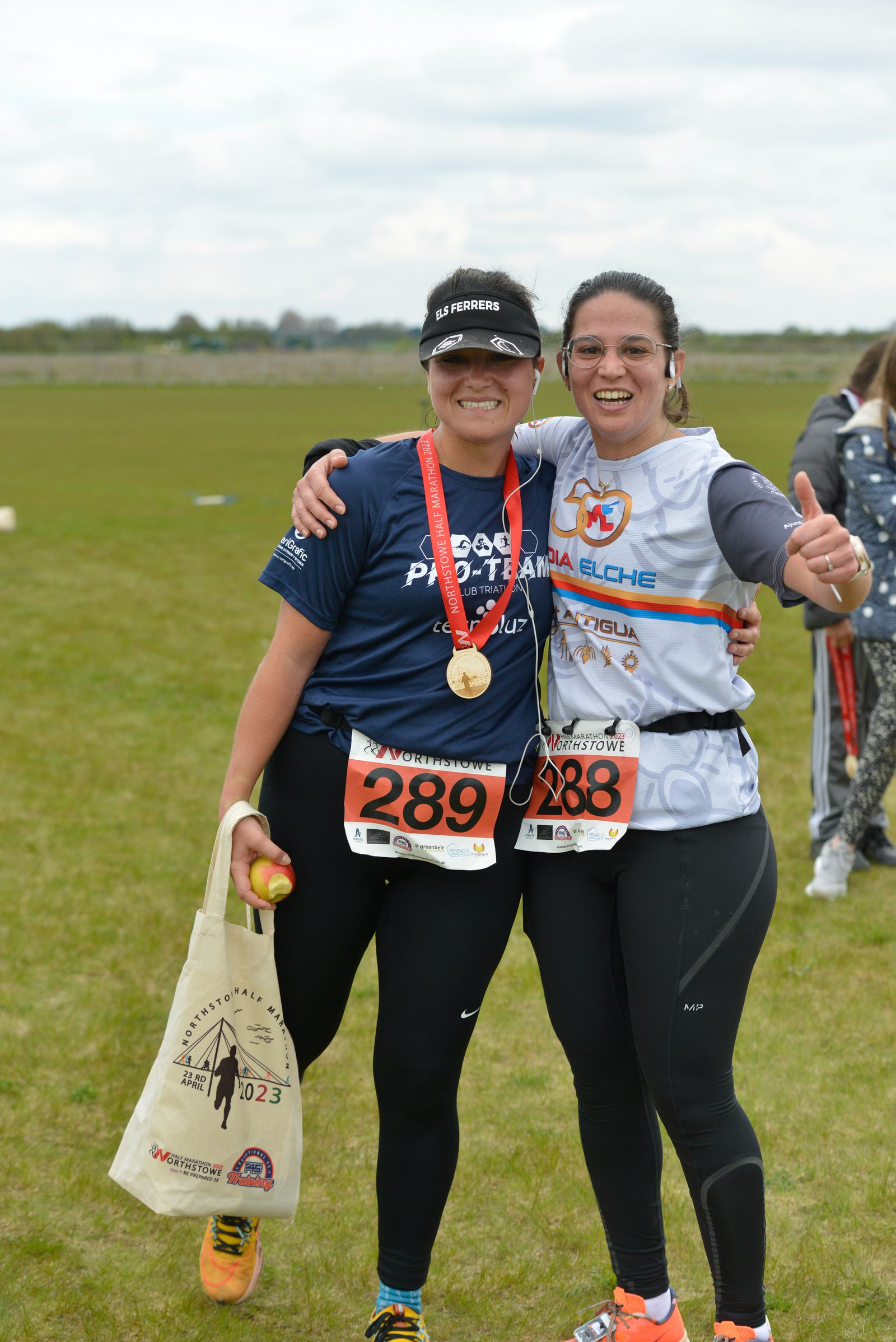 Two runners, both wearing numbered bibs and medals, pose together and smile after a race. The runner on the left carries a tote bag, while the one on the right gives a thumbs-up. They stand on a grassy field with other participants in the background.