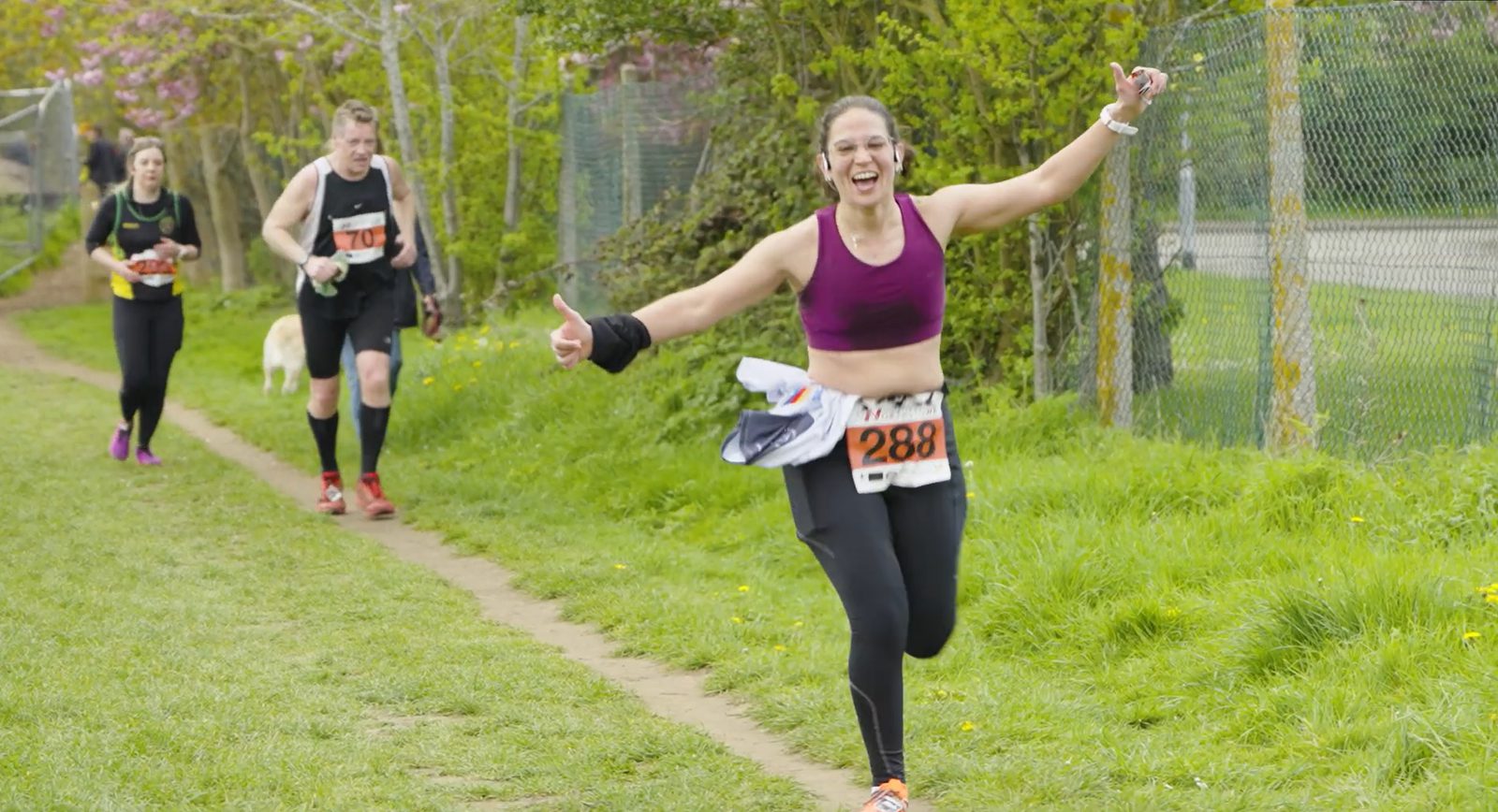 A woman in athletic gear, wearing a bib number 288, runs on a grassy path with her arms outstretched and a joyful expression. Behind her, other runners follow, one with a dog. The background includes greenery and a fence.