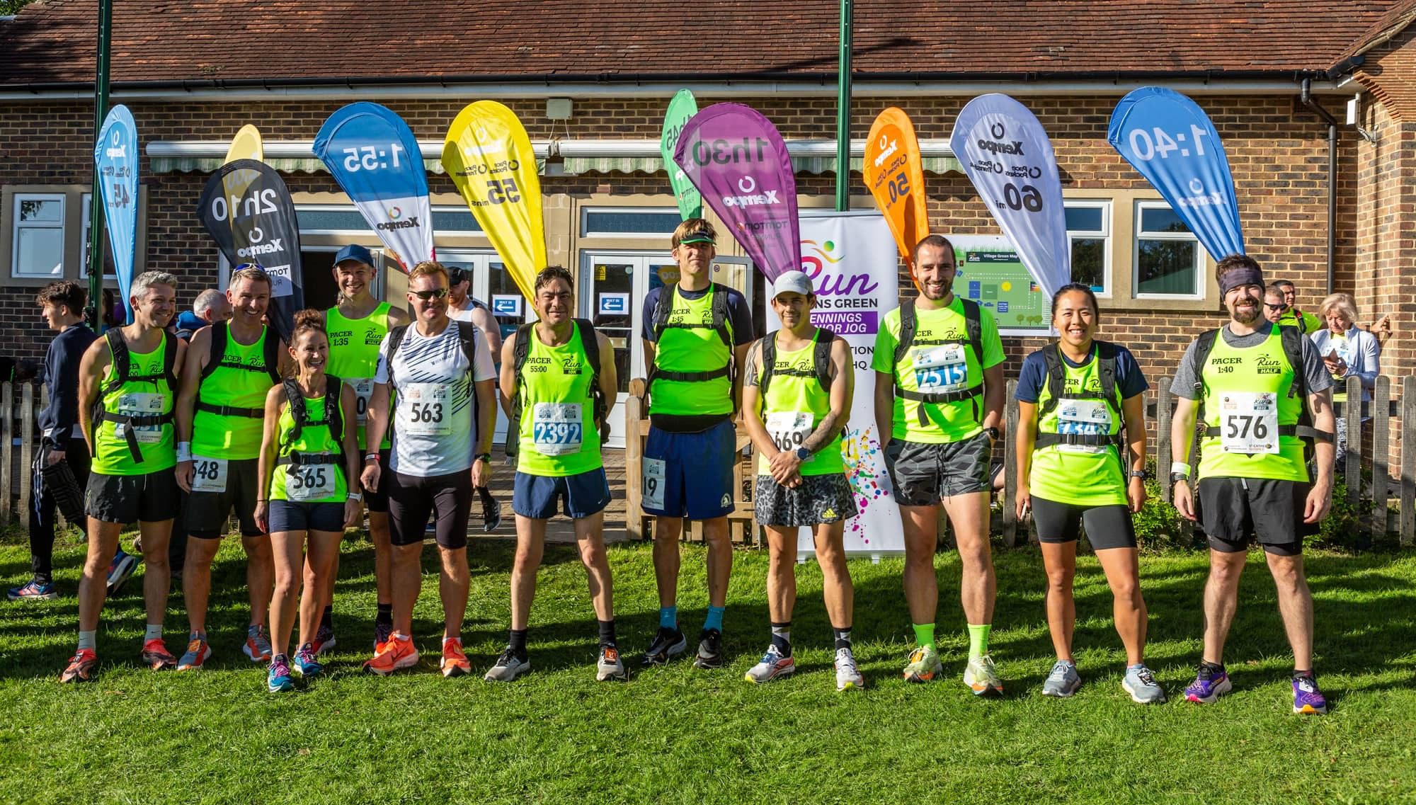 A group of diverse runners in green athletic gear pose for a photo outside a brick building with colorful banners behind them. They all have race bibs with numbers on their chests and are standing on a grassy area, smiling and ready for the event.