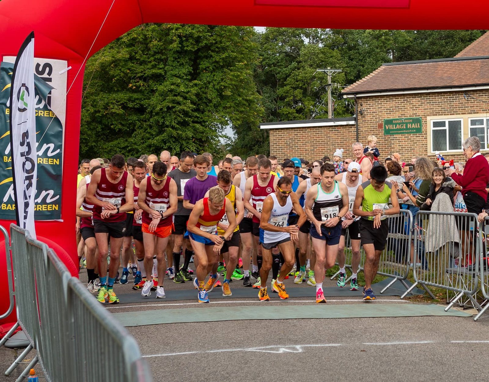 Runners in athletic gear gather at the starting line under a large red inflatable arch for a race. The course is flanked by metal barriers and a crowd of spectators. Some participants are crouched, ready to dash forward. A village hall building is visible in the background.