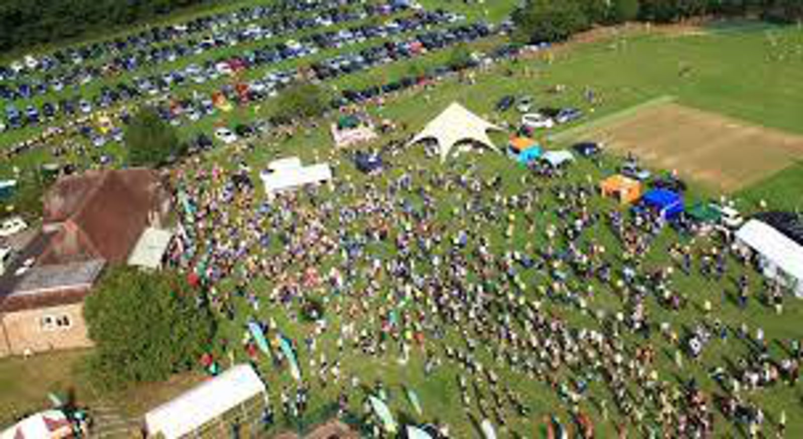 Aerial view of a large outdoor event with numerous people gathered on a grassy area. Various tents and booths are set up, along with a stage. Many cars are parked in an adjacent lot, and the surroundings are lined with trees.