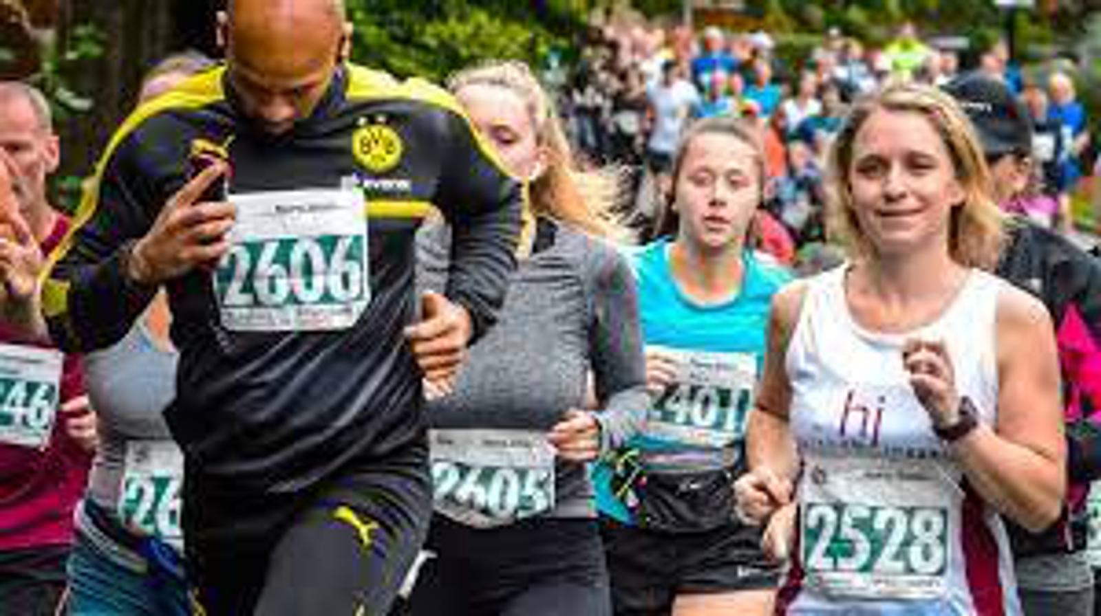 A group of runners participate in a road race. The central runner on the left wears a black and yellow outfit with the number 2606, while the runner on the right wears a white tank top with the number 2528. The background features more runners and a green, leafy environment.