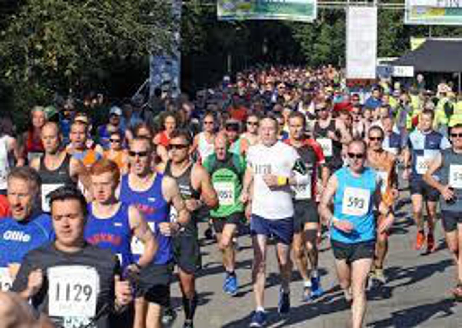 A large group of runners is participating in a race on a sunny day. They are wearing a variety of colorful athletic outfits and bib numbers. The race course is lined with trees, and spectators can be seen in the background.