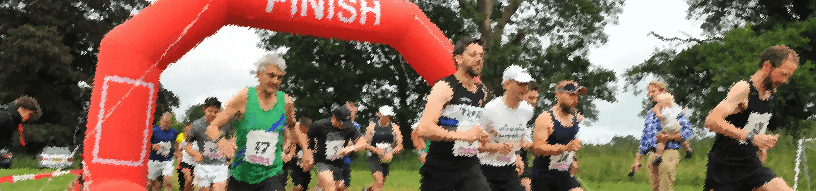 A group of runners, wearing race bibs, is seen crossing a red inflatable finish line arch in an outdoor marathon or race. Trees and grass are in the background, indicating the event takes place in a park or natural setting. The atmosphere appears energetic and enthusiastic.