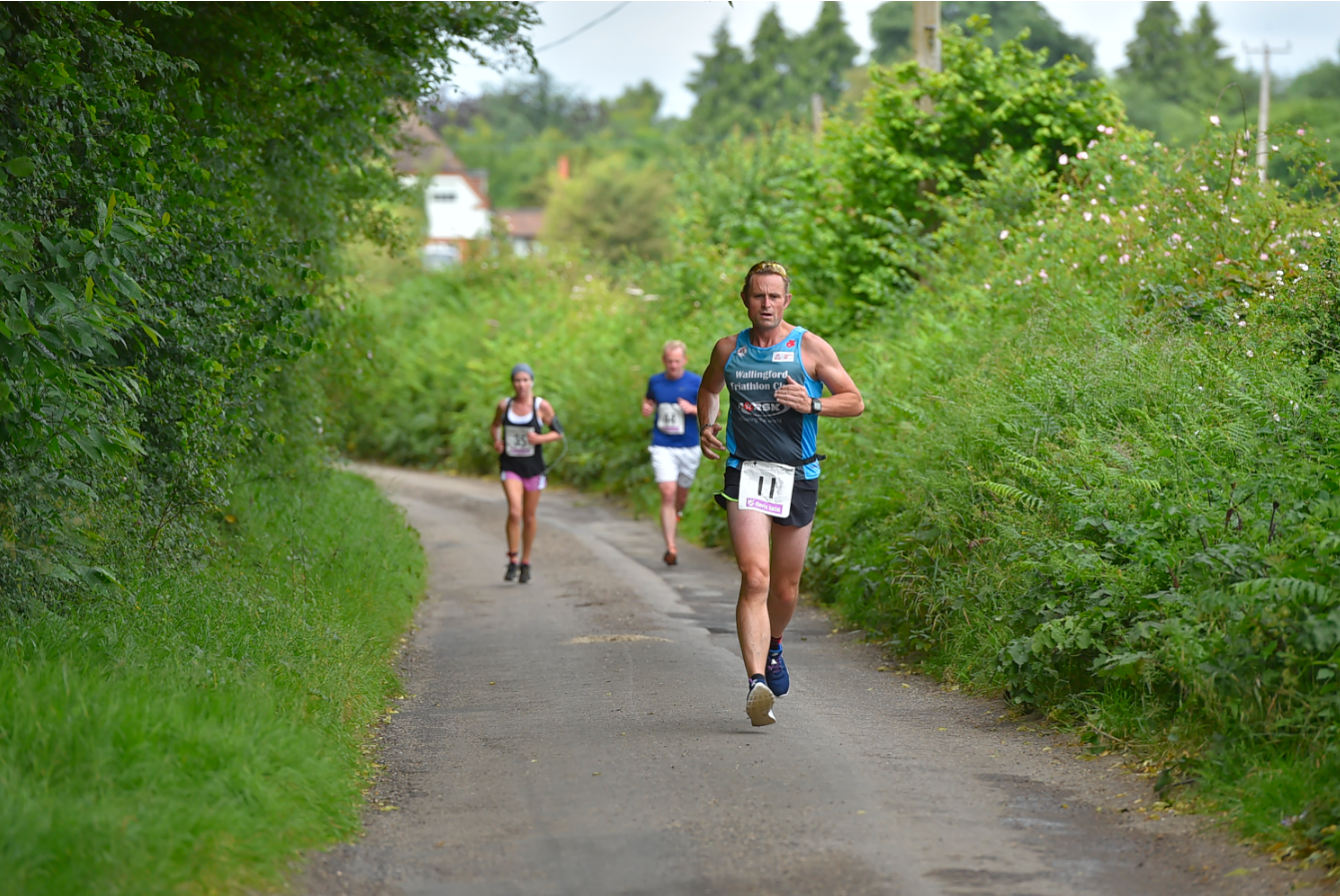 A male runner in a green singlet and black shorts with a race bib number 11 jogs along a narrow, tree-lined country path. Three other runners are visible in the background. The sky is partly cloudy, and the lush green surroundings suggest it's a summer day.