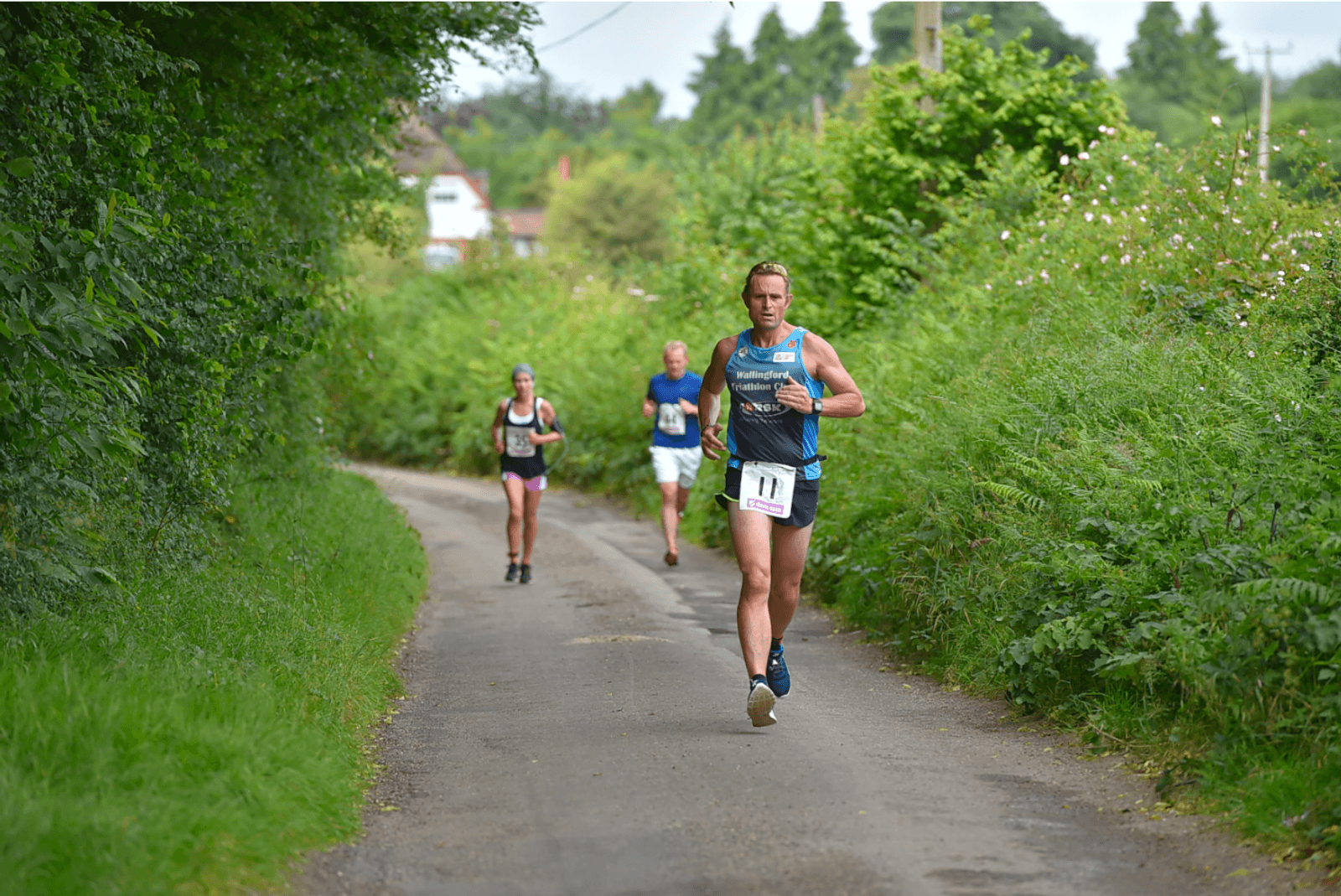 A male runner in a green singlet and black shorts with a race bib number 11 jogs along a narrow, tree-lined country path. Three other runners are visible in the background. The sky is partly cloudy, and the lush green surroundings suggest it's a summer day.