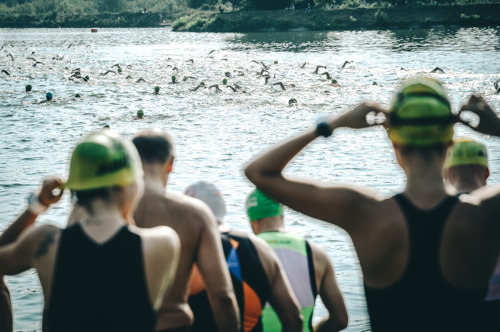 Photo of participants in a triathlon swim event. Swimmers wearing swim caps and wetsuits head towards a body of water. Some are in the water, while others adjust their gear on the shore, preparing to start. Trees and greenery line the background.