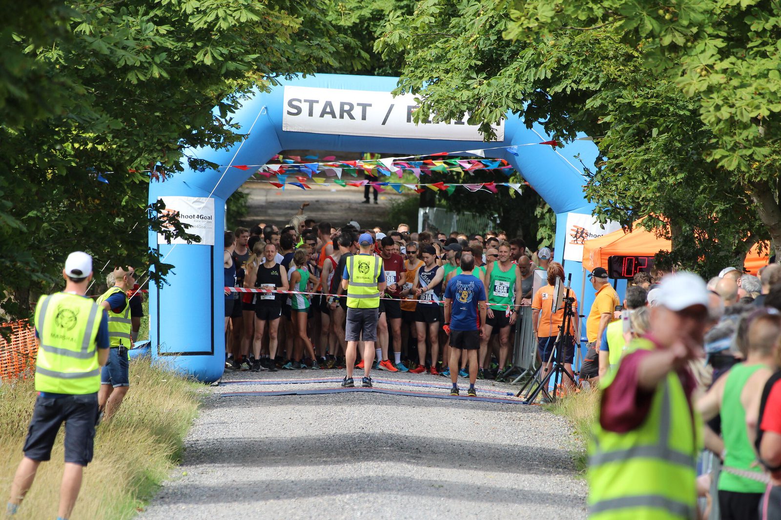 A large group of runners gathers at the starting line under a blue inflatable arch with "START" written on it. Trees line both sides, providing shade. People in yellow vests are seen managing the event, and colorful flags hang above the path.