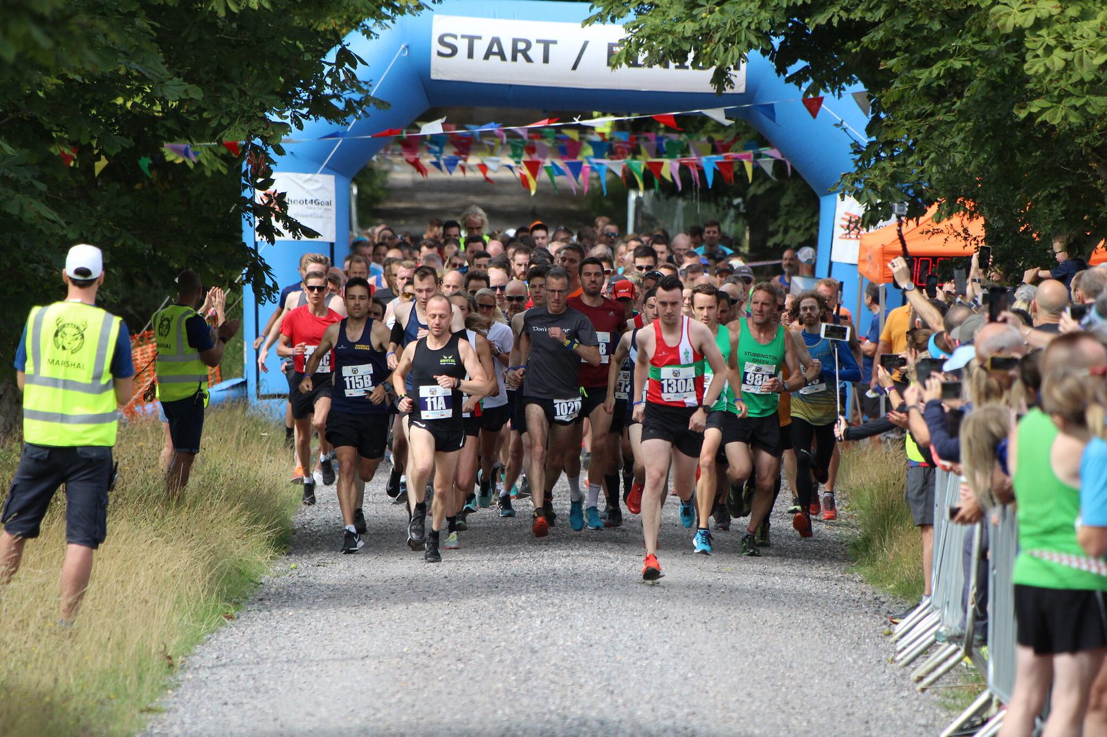 A group of runners, both men and women, are starting a race on a gravel path. They are passing under a blue inflatable arch with "START" written on it. Spectators stand on both sides of the path, and colorful flags are strung above the arch.