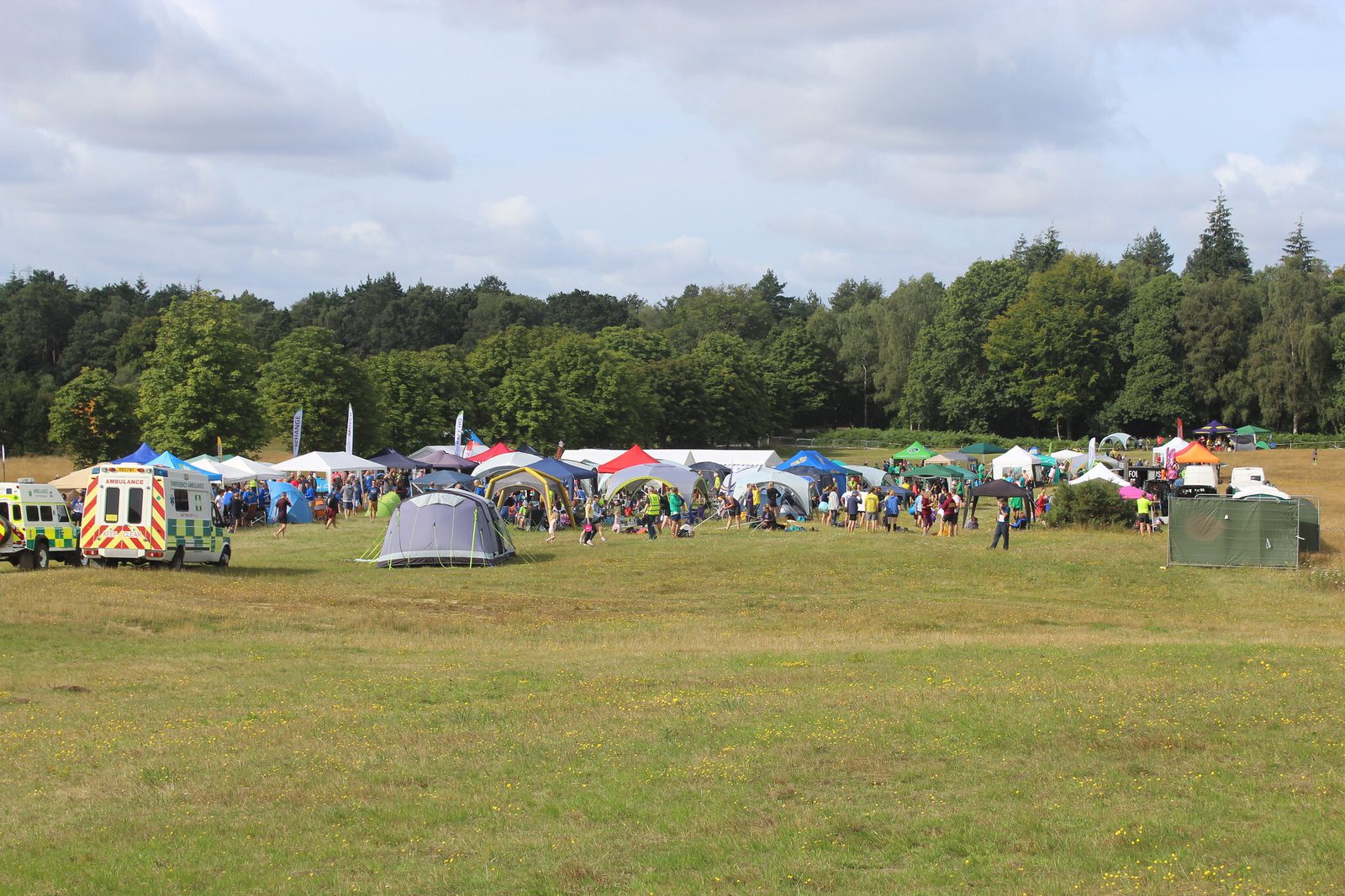 A large outdoor gathering on a grassy field with numerous tents and canopies set up. People walk around and socialize. An ambulance and a vehicle are visible on the left, while trees and a blue sky are in the background.