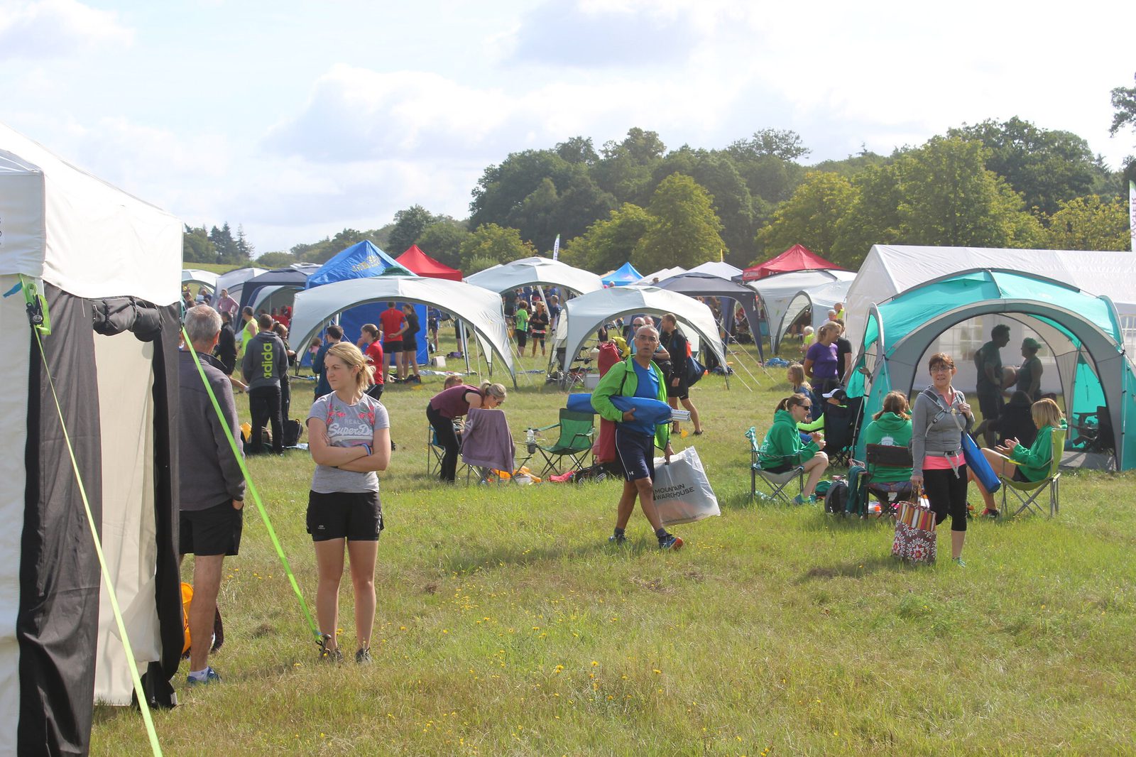 A busy campsite scene with various tents and canopies set up in a grassy field. People are walking, sitting on camp chairs, and socializing. The weather is sunny with a few clouds, and green trees are visible in the background.