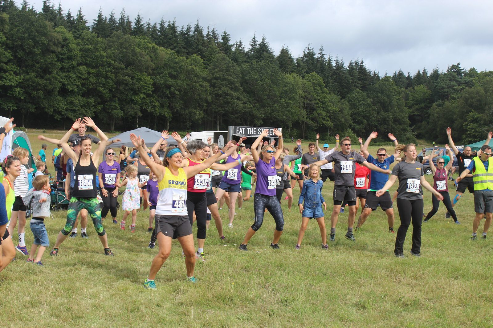 A large group of people exercising outdoors in a grassy area, many of them raising their arms. They are of different ages, wearing fitness attire. A food truck and dense green trees are visible in the background. Some children and spectators are also present.