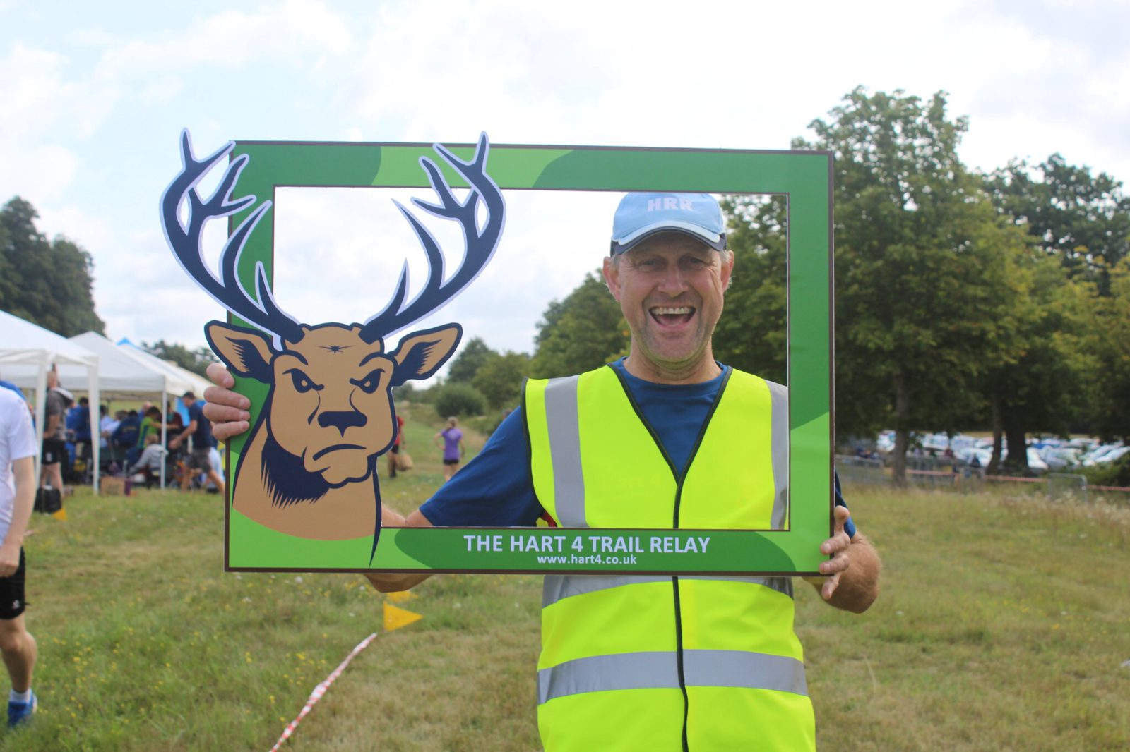 A cheerful man wearing a high-visibility vest and a blue cap is holding a large green frame with an illustrated deer head on it and "The Hart 4 Trail Relay" text. He is standing outdoors in a grassy area with trees and tents in the background.