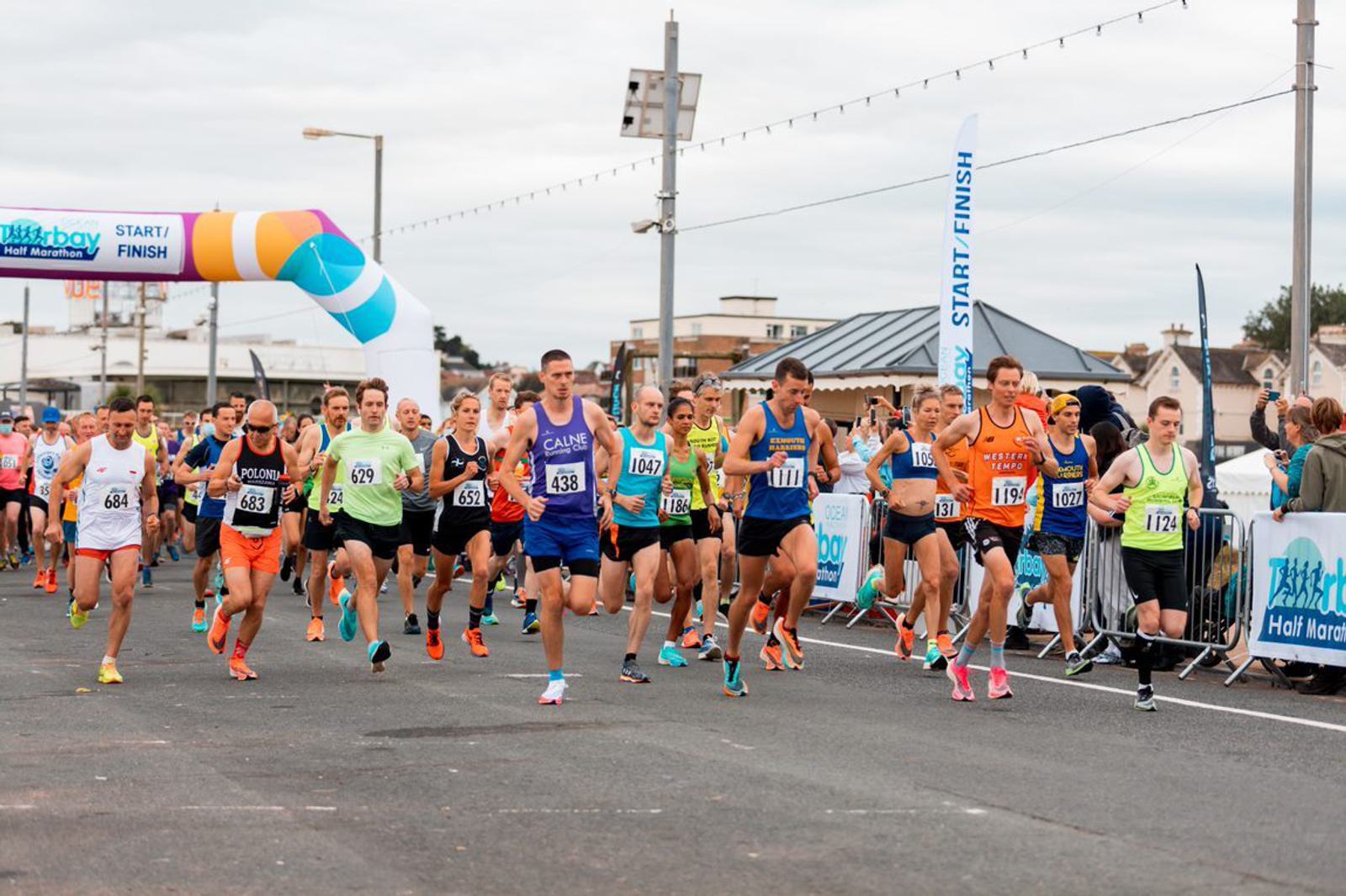 A group of runners starting a race at the Halfway 10K event. Participants are wearing running gear with various bib numbers and are running down a paved road. A colorful archway and "START/FINISH" signs are visible in the background, along with spectators.