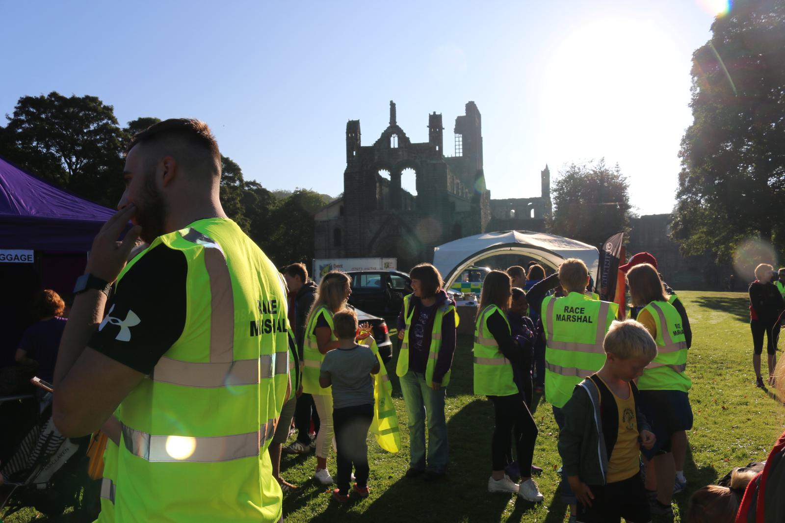 A group of people wearing yellow vests, some labeled "RACE MARSHAL," gather at an outdoor event on a sunny day. A historic stone building and trees are visible in the background. A few children are present, and a large tent is set up nearby.