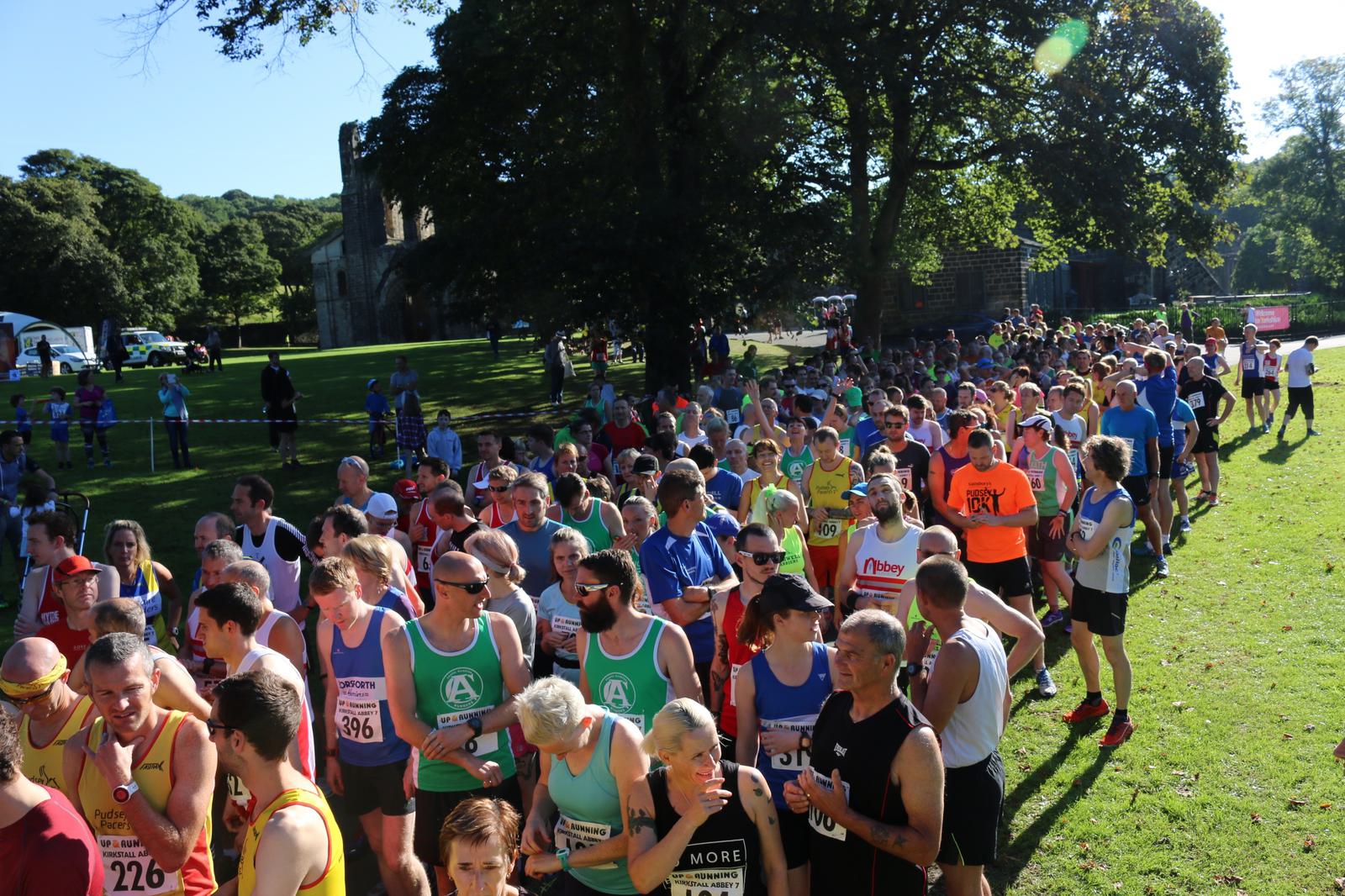A large group of runners in various colorful athletic outfits gather at the starting line of an outdoor race event. The scene is set in a lush, green park on a sunny day with trees and some structures visible in the background.