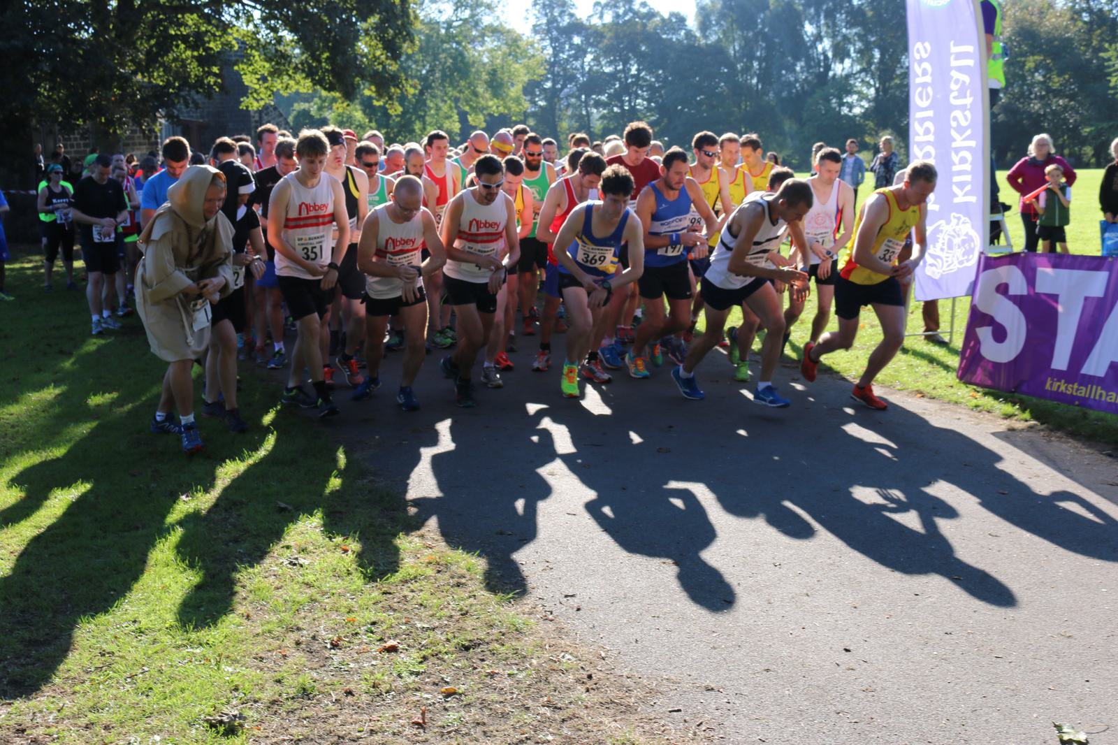 A group of runners wearing athletic gear are starting a race on a sunny day. Some runners are wearing bibs with numbers. Trees and grass surround the starting area, and there is a purple race banner on the right side of the image.